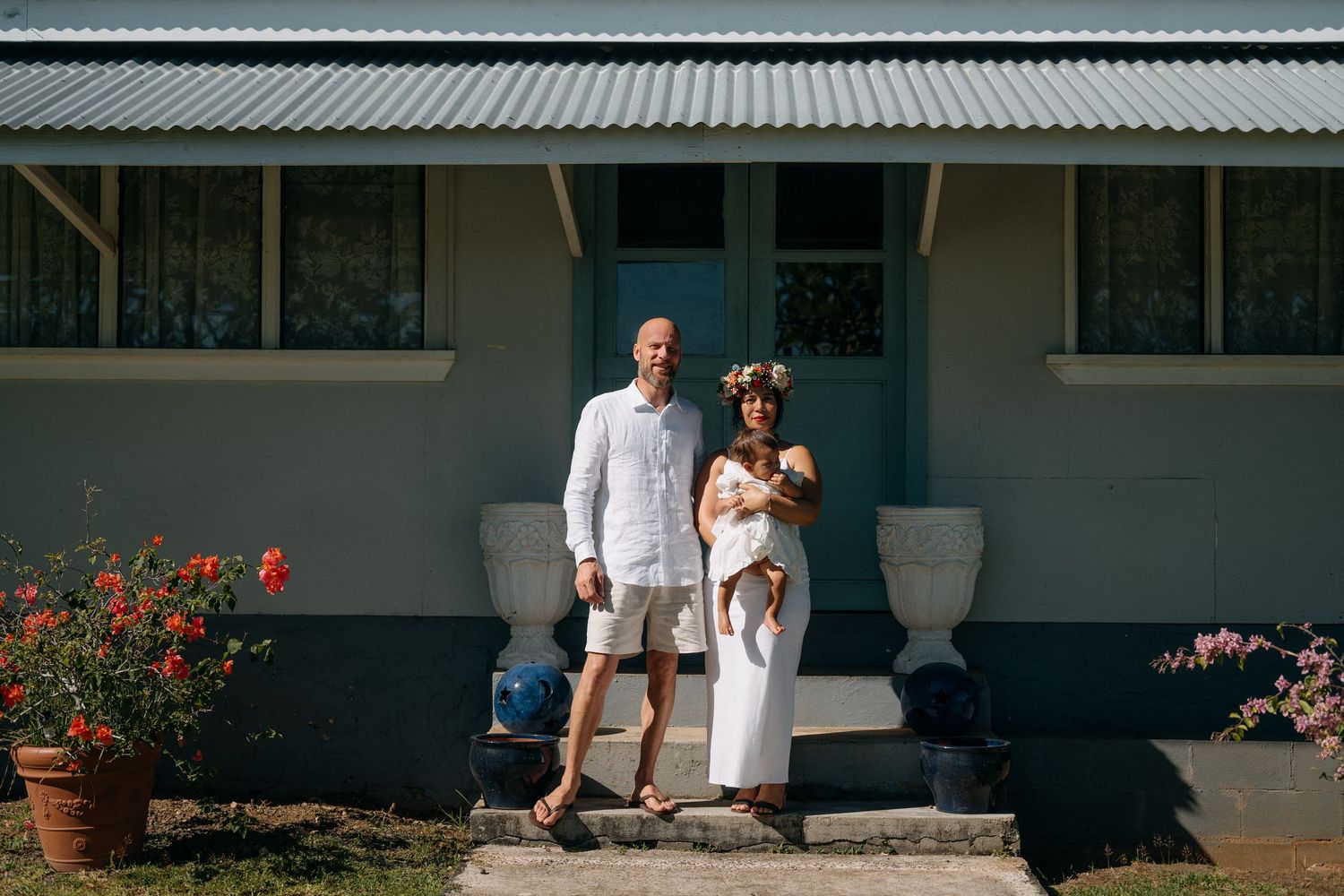 A couple with a baby stands on steps of a light blue house, woman wearing floral crown and white dress.