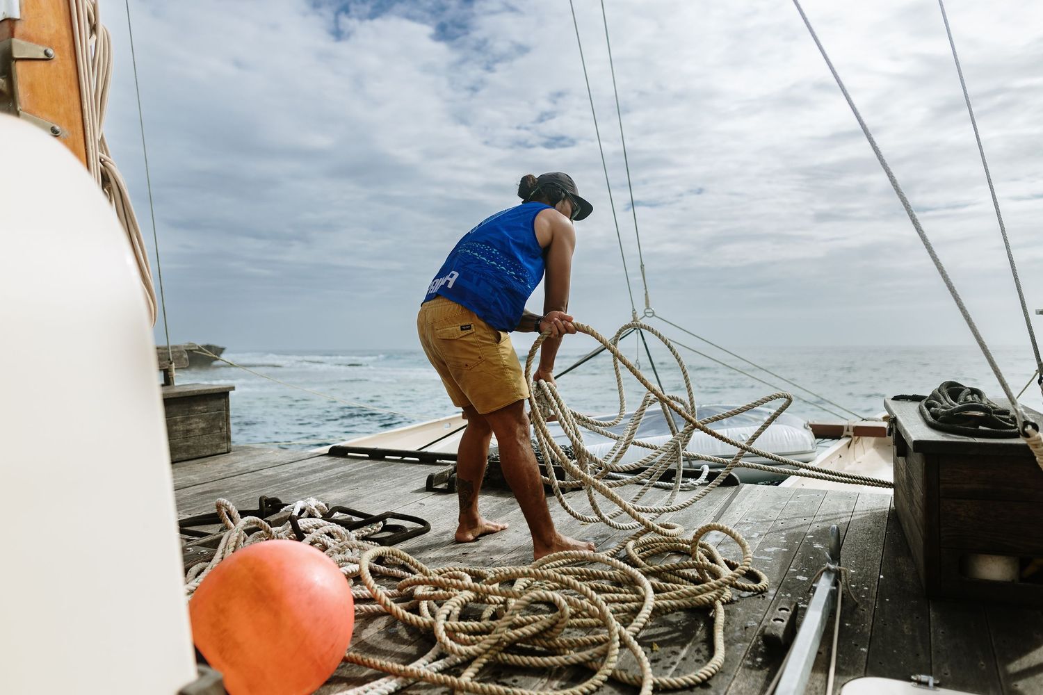 Person coiling rope on sailboat deck with ocean backdrop, wearing blue shirt and tan shorts.