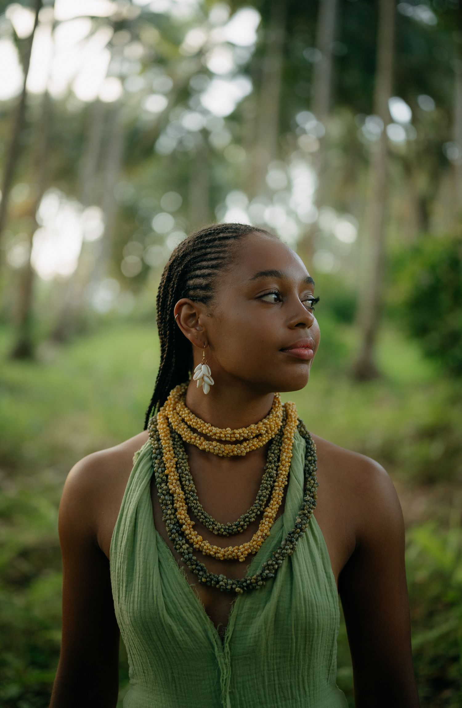 Young Black woman with braids wearing layered beaded necklaces and green dress standing in lush tropical forest.