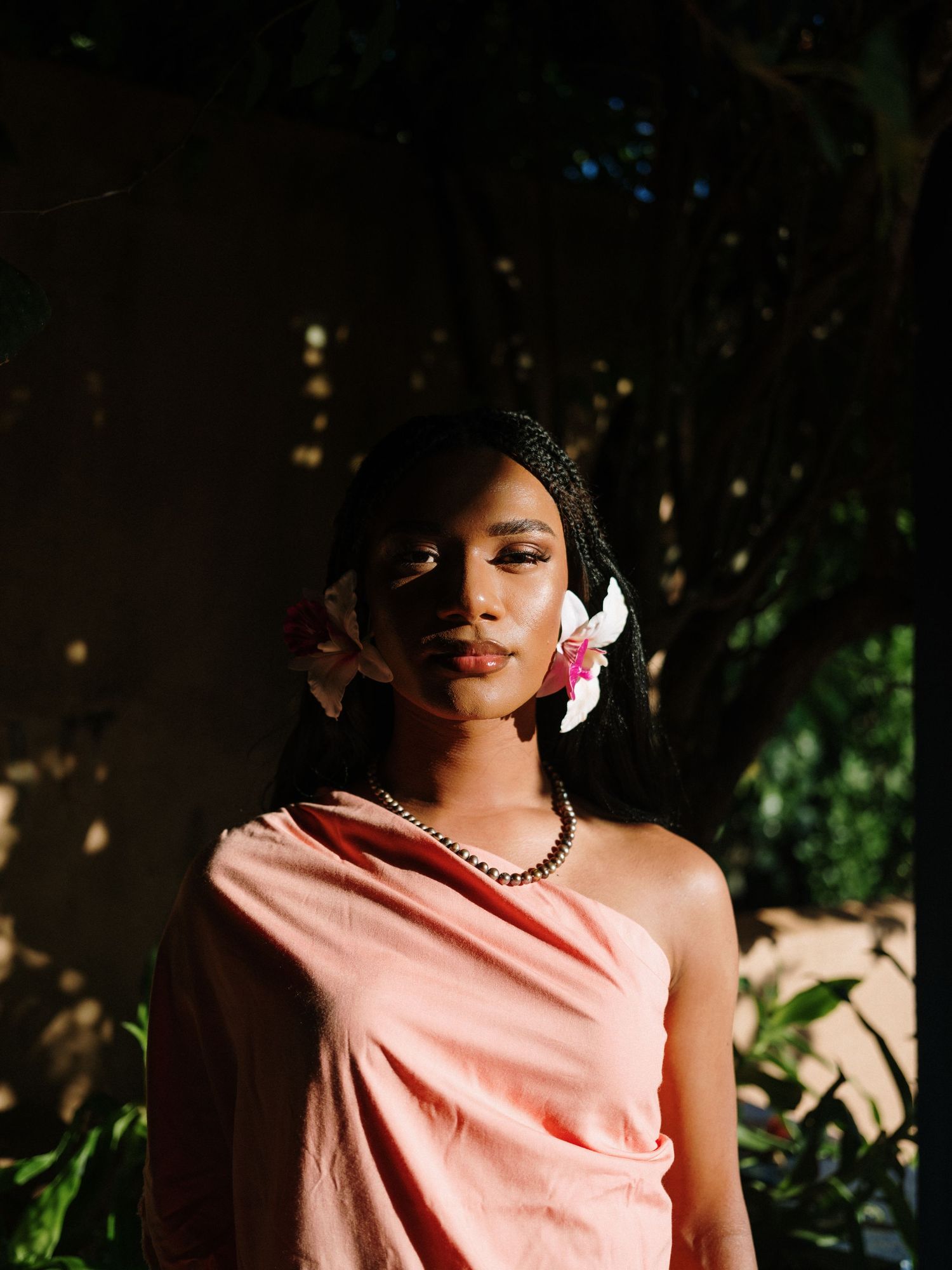 Young woman in pink draped dress with flowers in braided hair, standing in dappled sunlight among lush greenery.