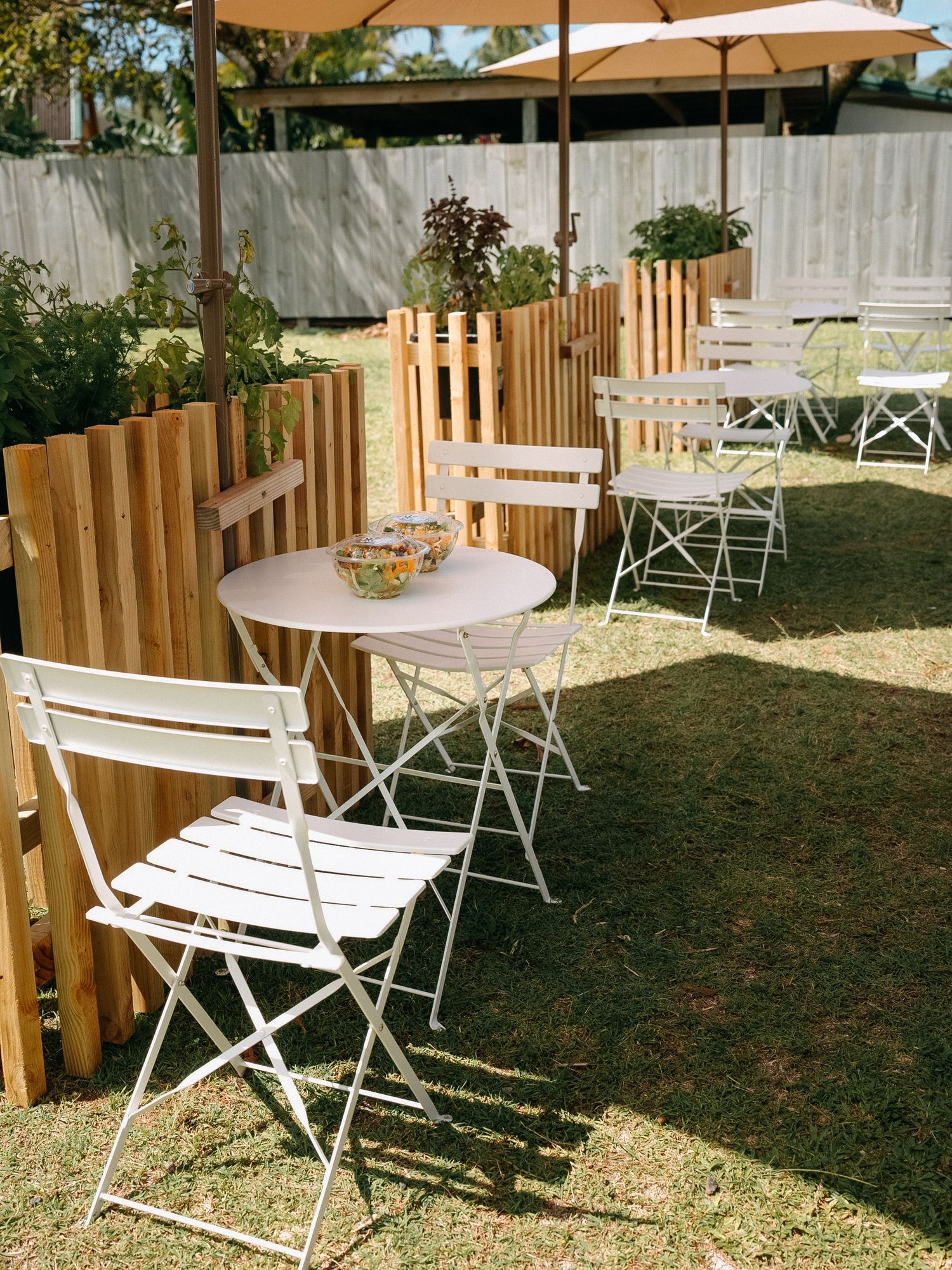 Outdoor cafe patio with white folding chairs, bistro tables, wooden fence, and patio umbrellas on green grass.