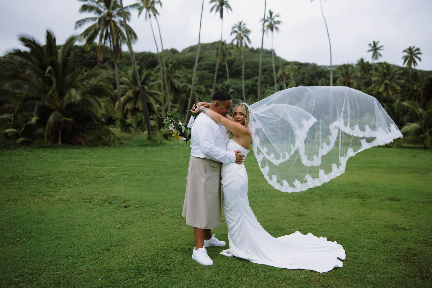 Couple embracing on lush green lawn with wedding veil flowing in wind, tropical palm trees in background.