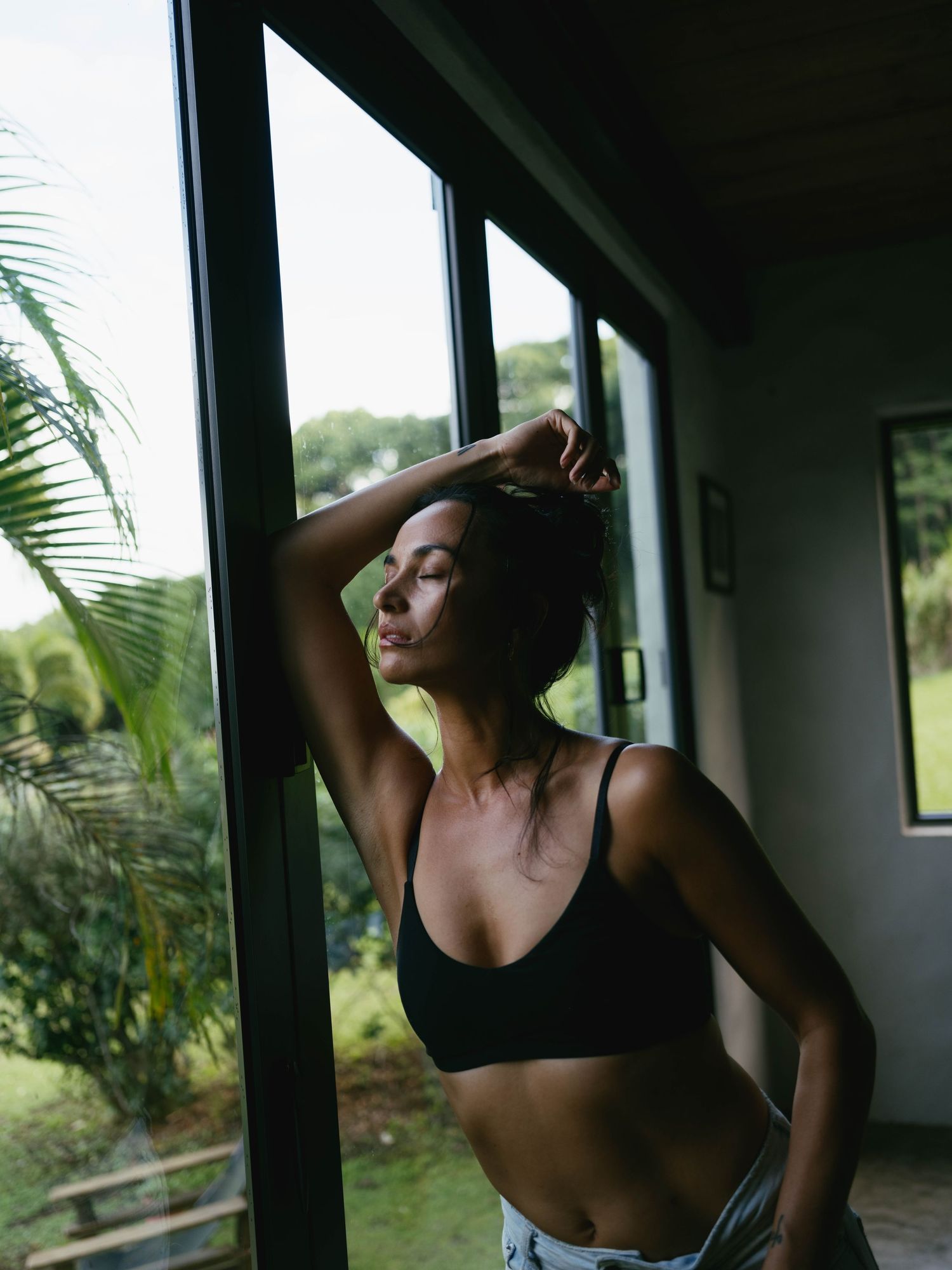 Woman in black sports bra stands by large window with tropical garden view, eyes closed, hand in hair.