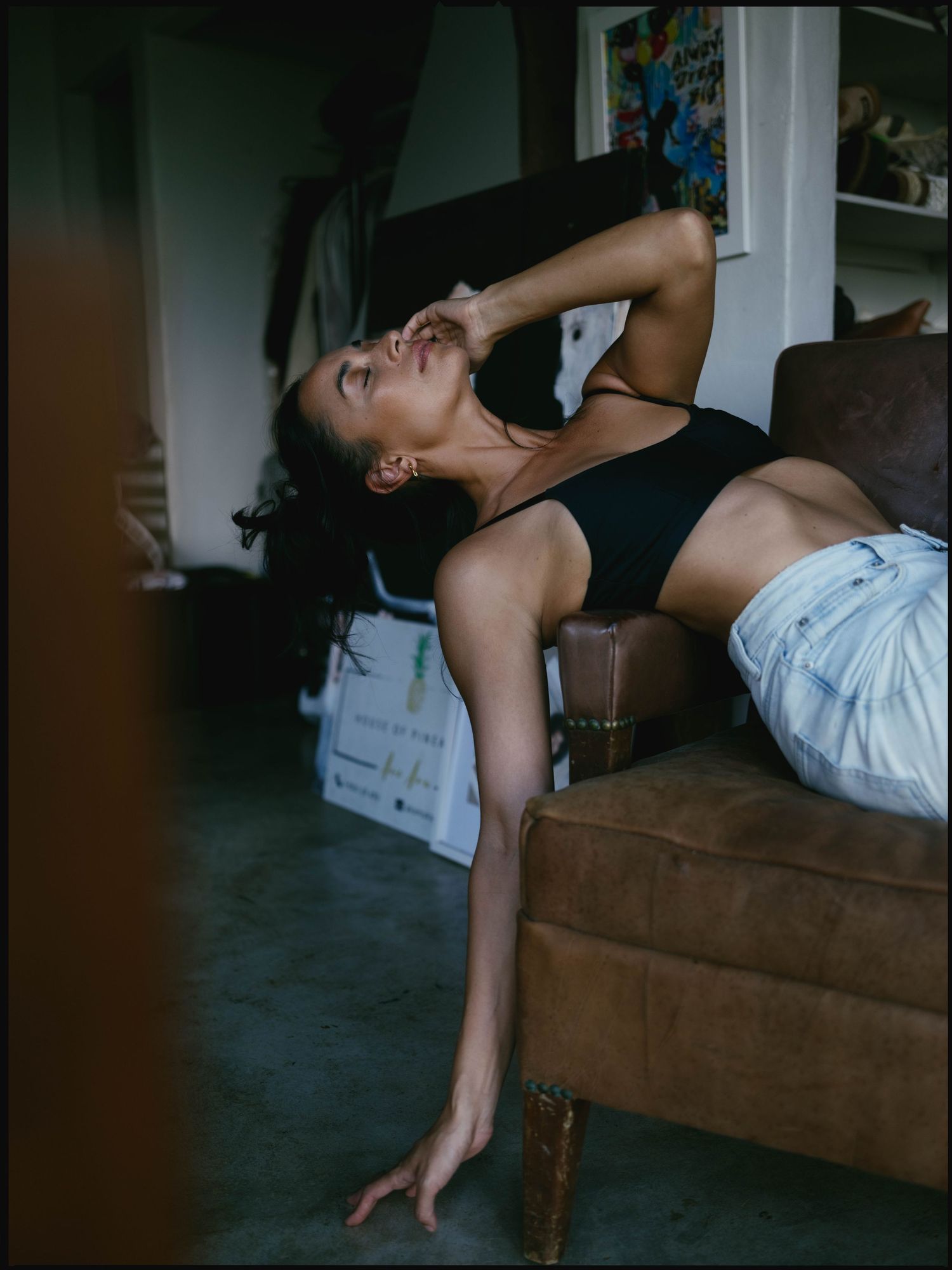 A woman in a black crop top and denim shorts reclines gracefully on a brown leather chair indoors.
