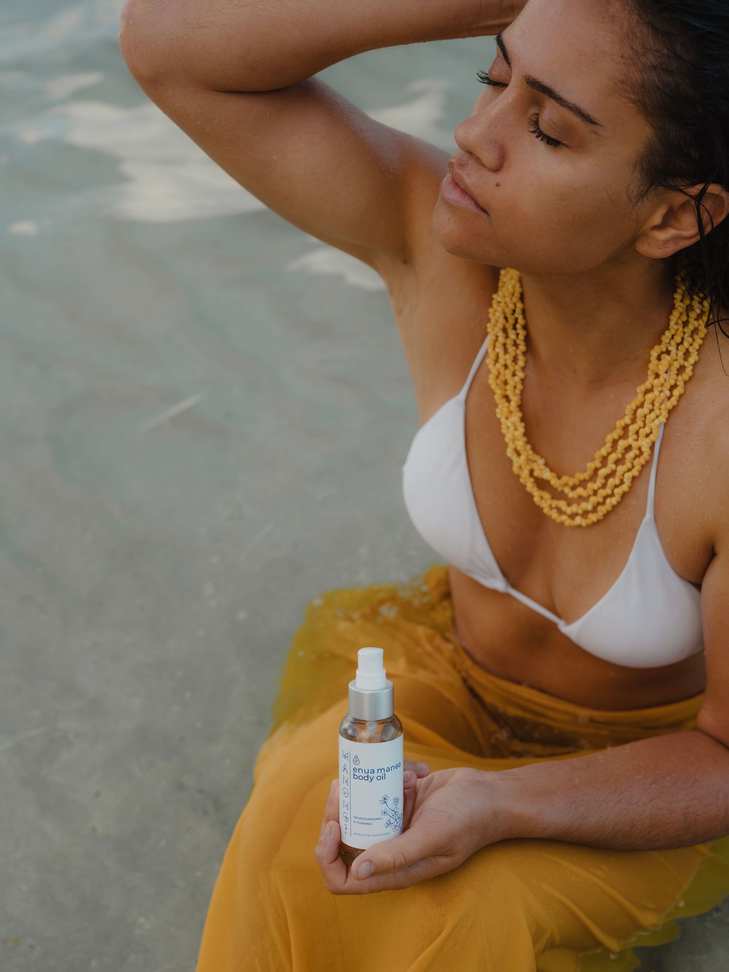 Woman in white bikini top holding a spray bottle near the beach, wearing a yellow beaded necklace.