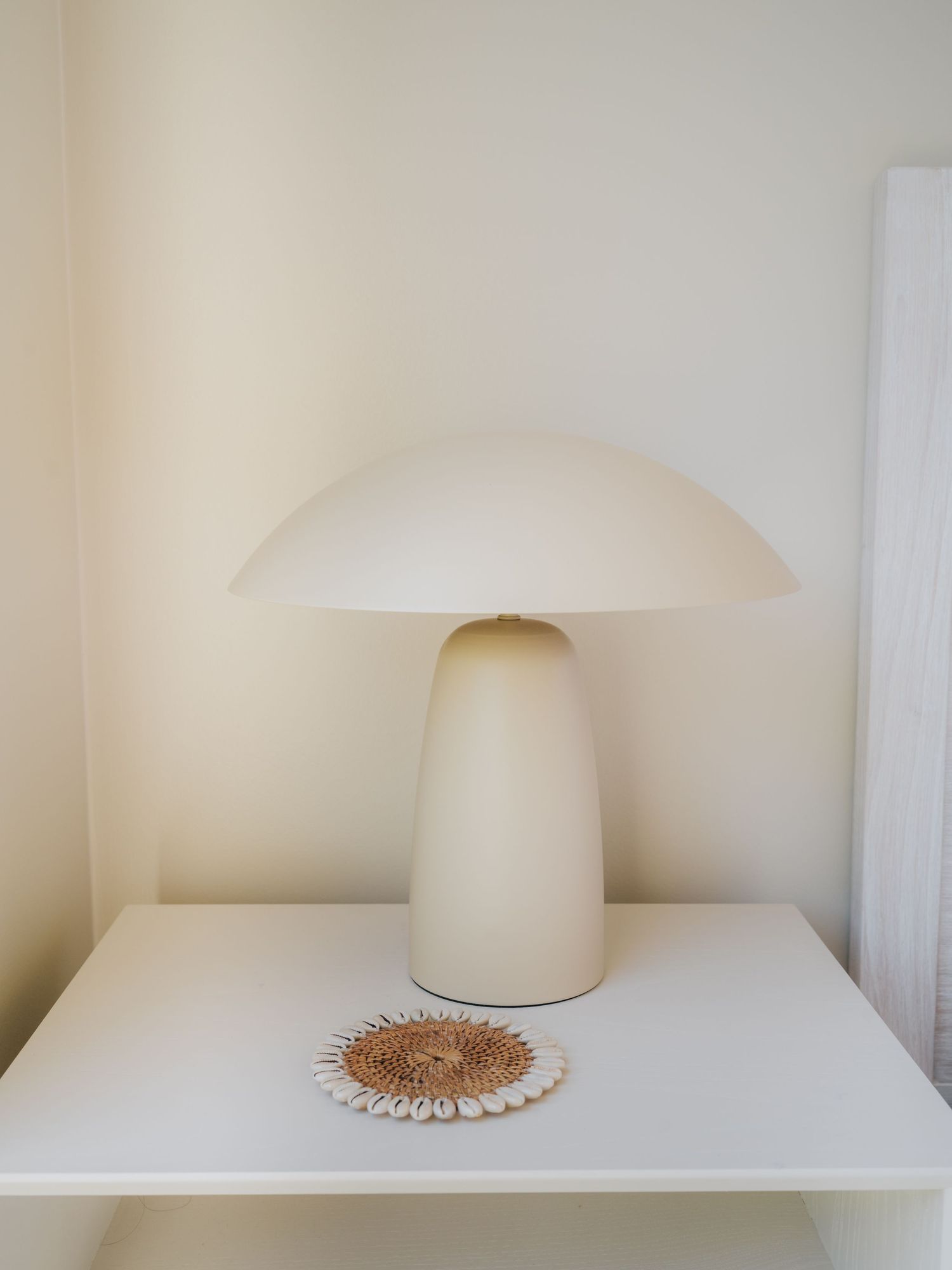 Cream mushroom-shaped table lamp on white shelf with woven rattan coaster and shell decorative trivet.