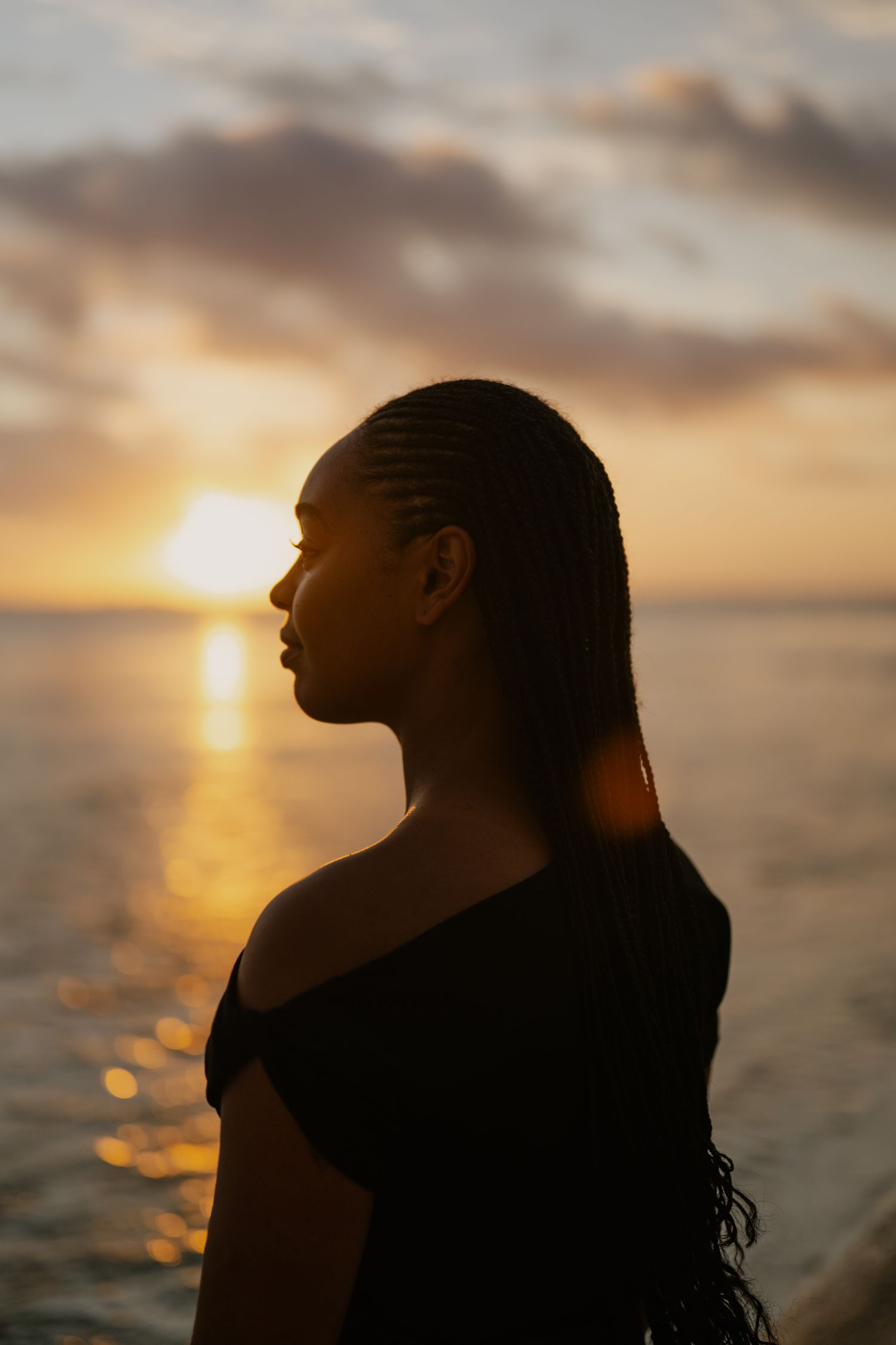 A young Black woman with braided hair stands in silhouette against a golden ocean sunset, wearing an off-shoulder black top.