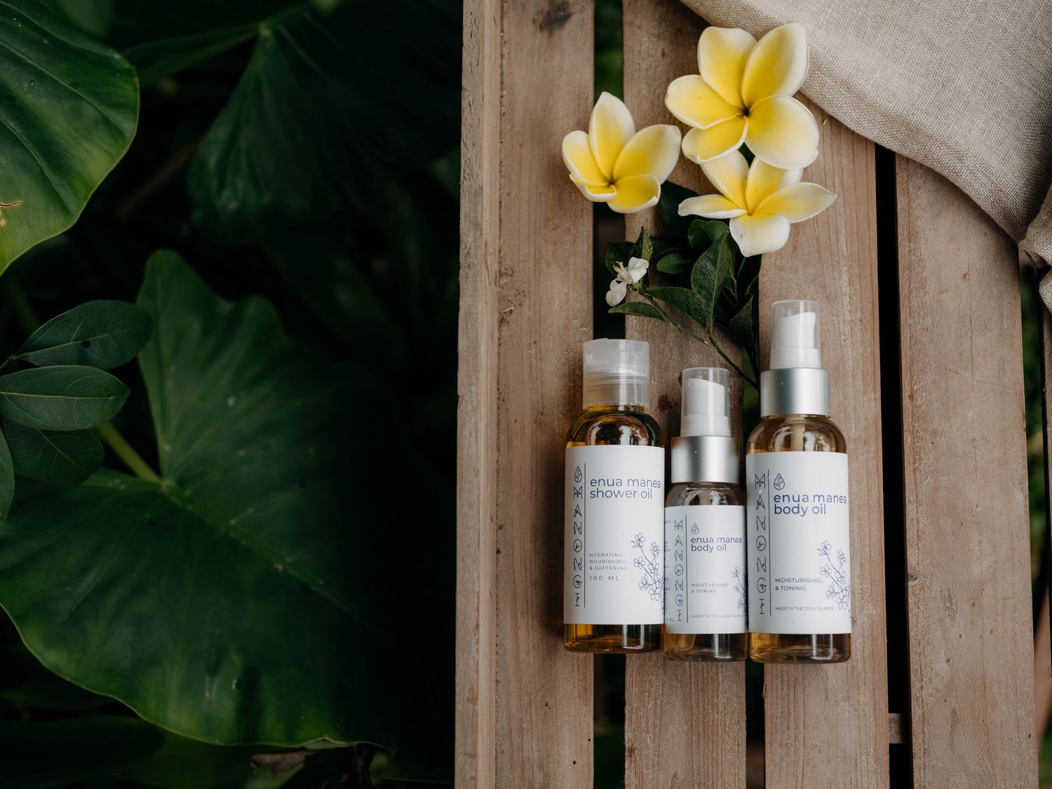 Three amber spray bottles with white labels beside yellow plumeria flowers on a wooden surface with tropical green leaves.