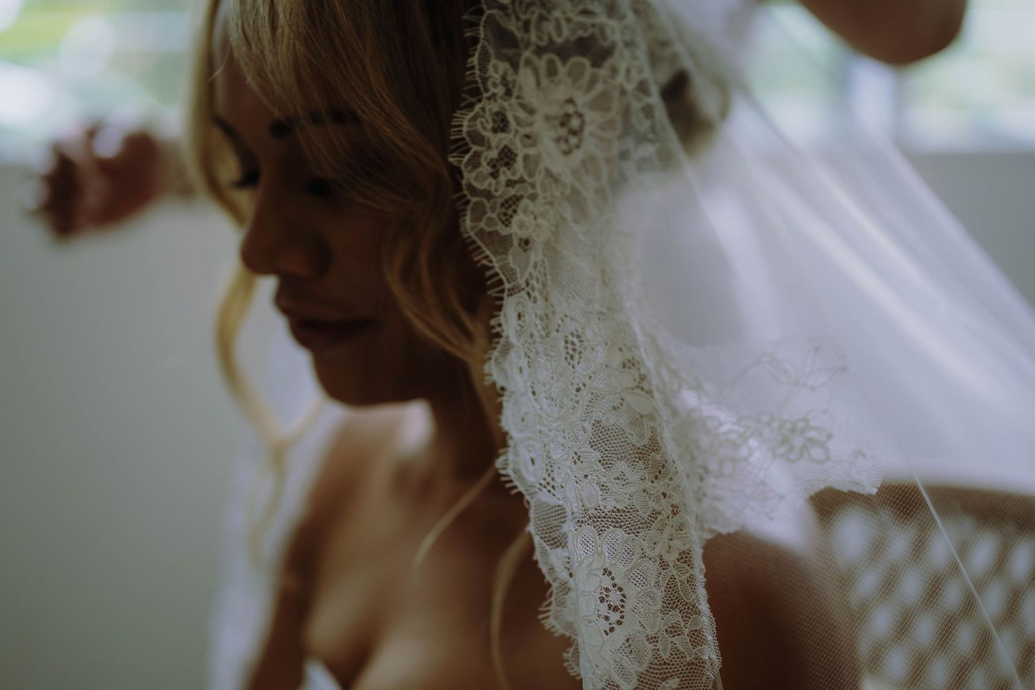 Bride wearing a delicate lace wedding veil with blonde hair, captured in a soft, moody close-up portrait.