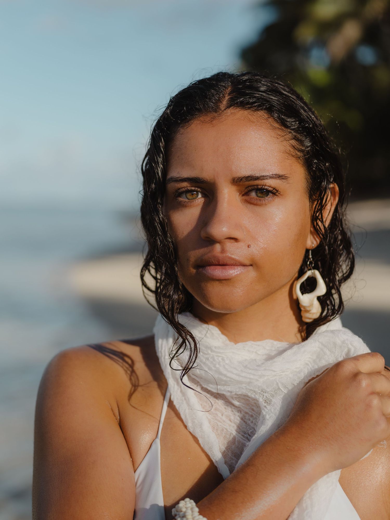 Young woman with wet hair and shell earrings wrapped in white fabric on a tropical beach.