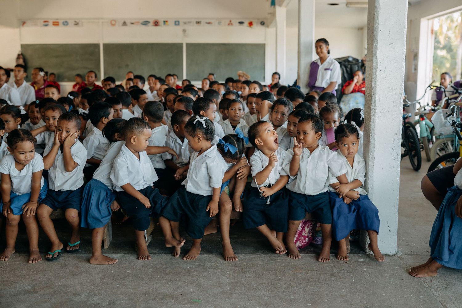 Large group of young students in school uniforms sitting together in an open-air classroom in Southeast Asia.