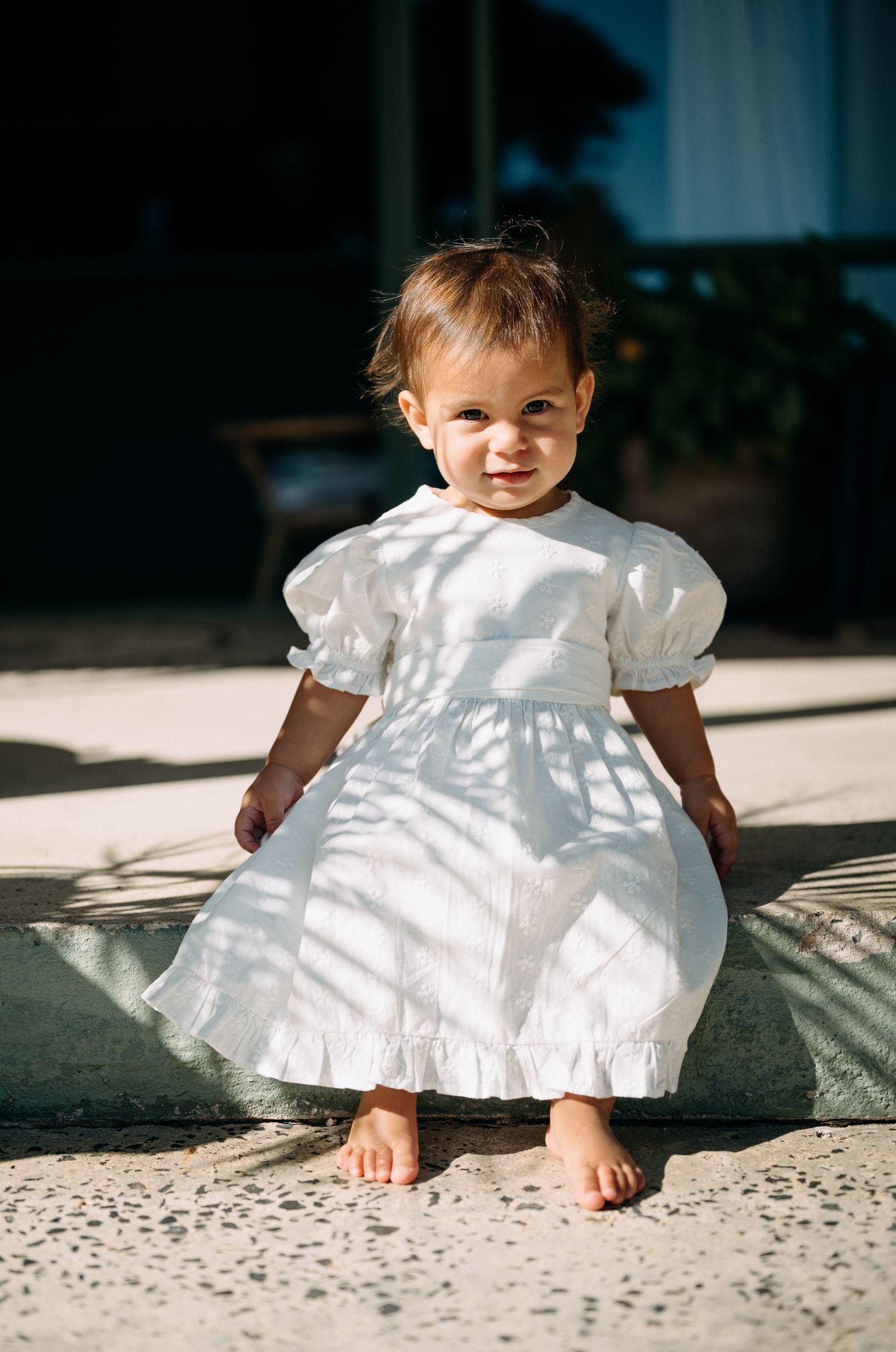 Adorable toddler girl in a white puff sleeve dress standing barefoot in sunlight with leaf shadows.