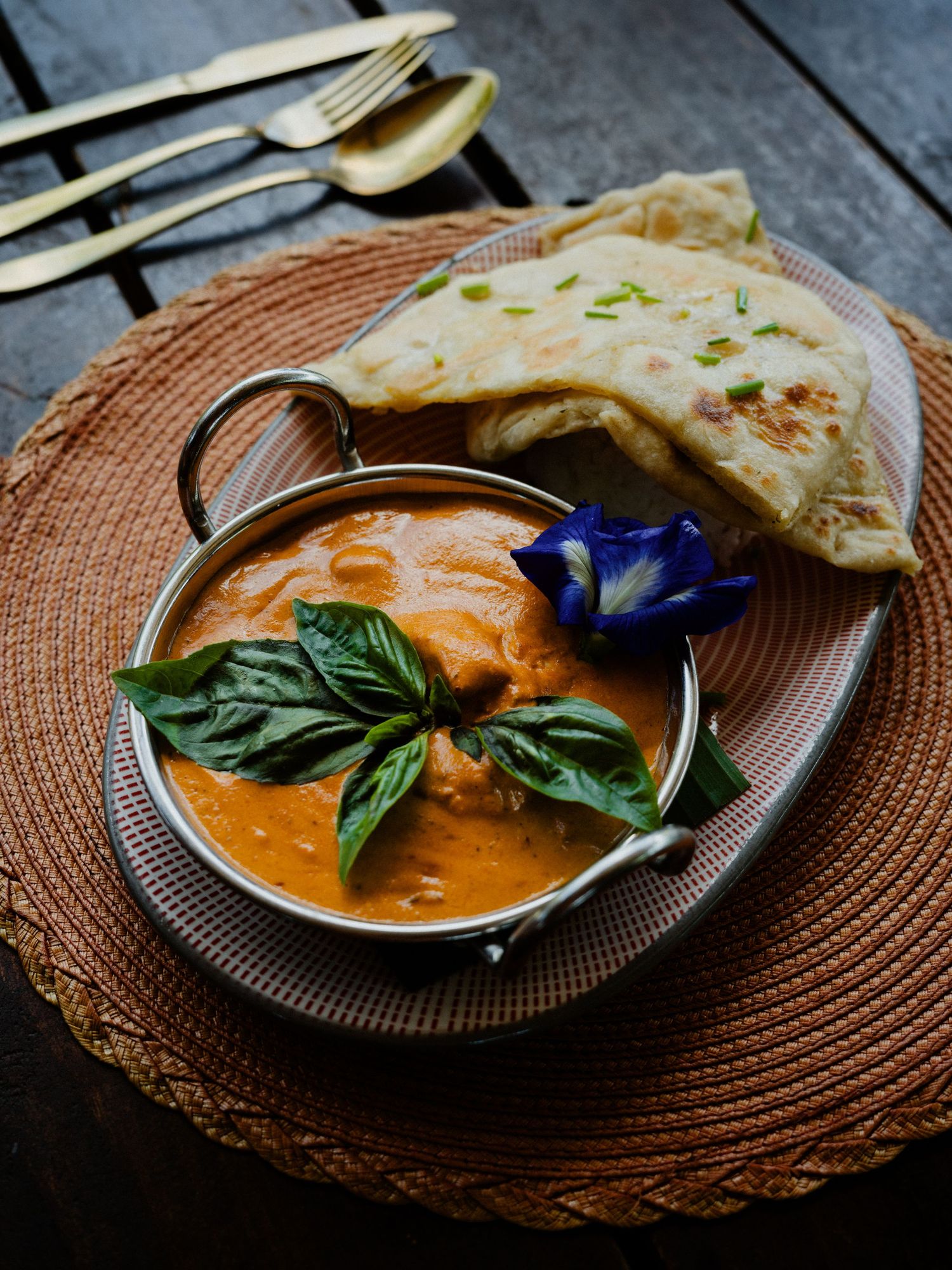 A rich butter chicken curry in a silver karahi bowl, garnished with basil and blue flower, served with naan bread.