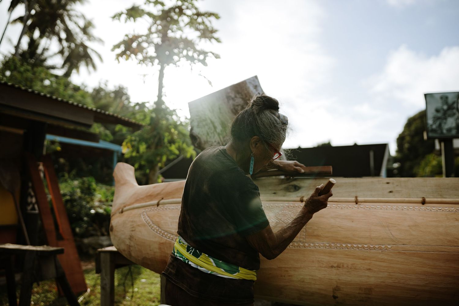 Craftsman carefully sanding a wooden canoe outdoors in a tropical setting with lush green trees in the background.
