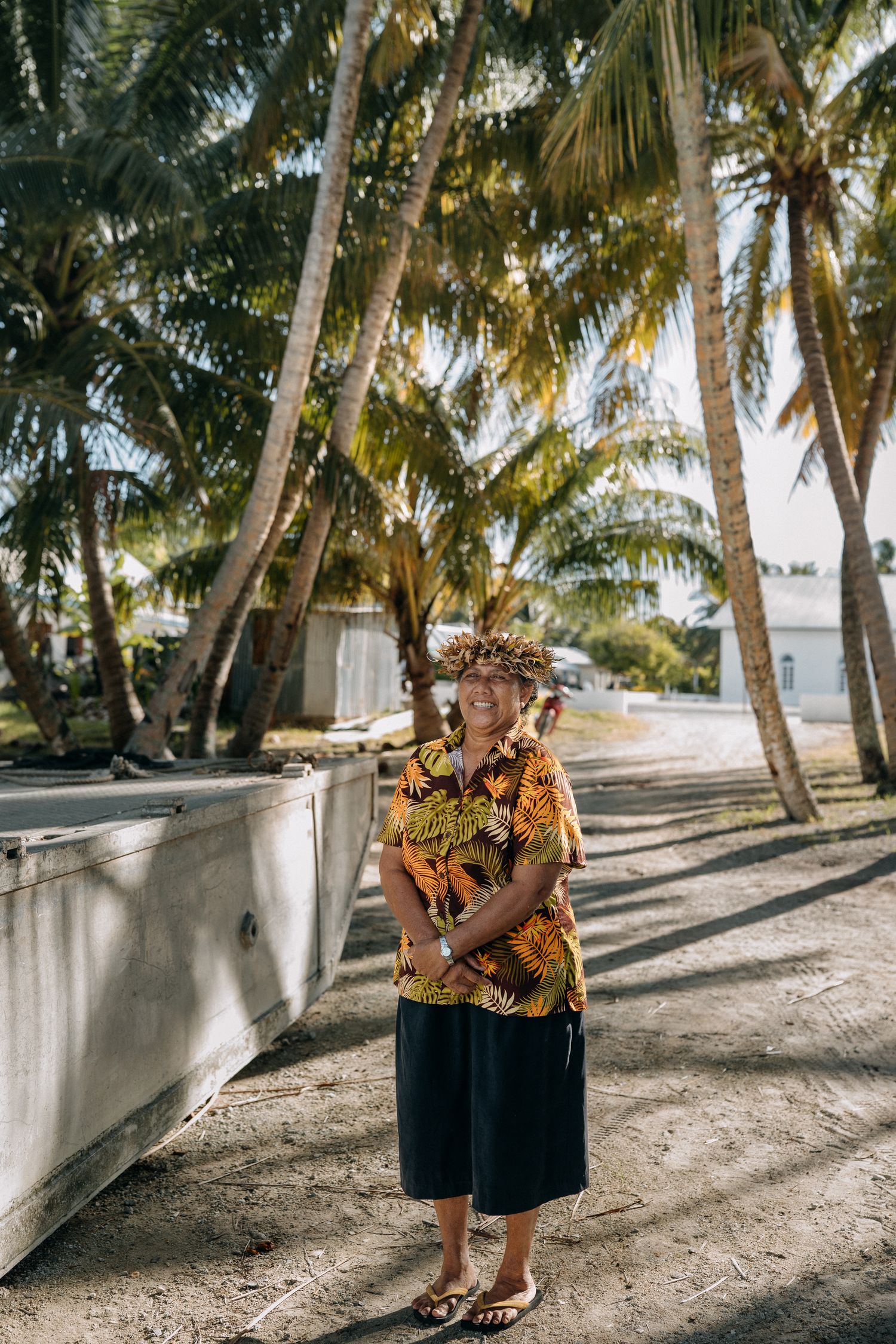 Smiling woman in floral shirt and flower crown stands among tropical palm trees on a sunny day.