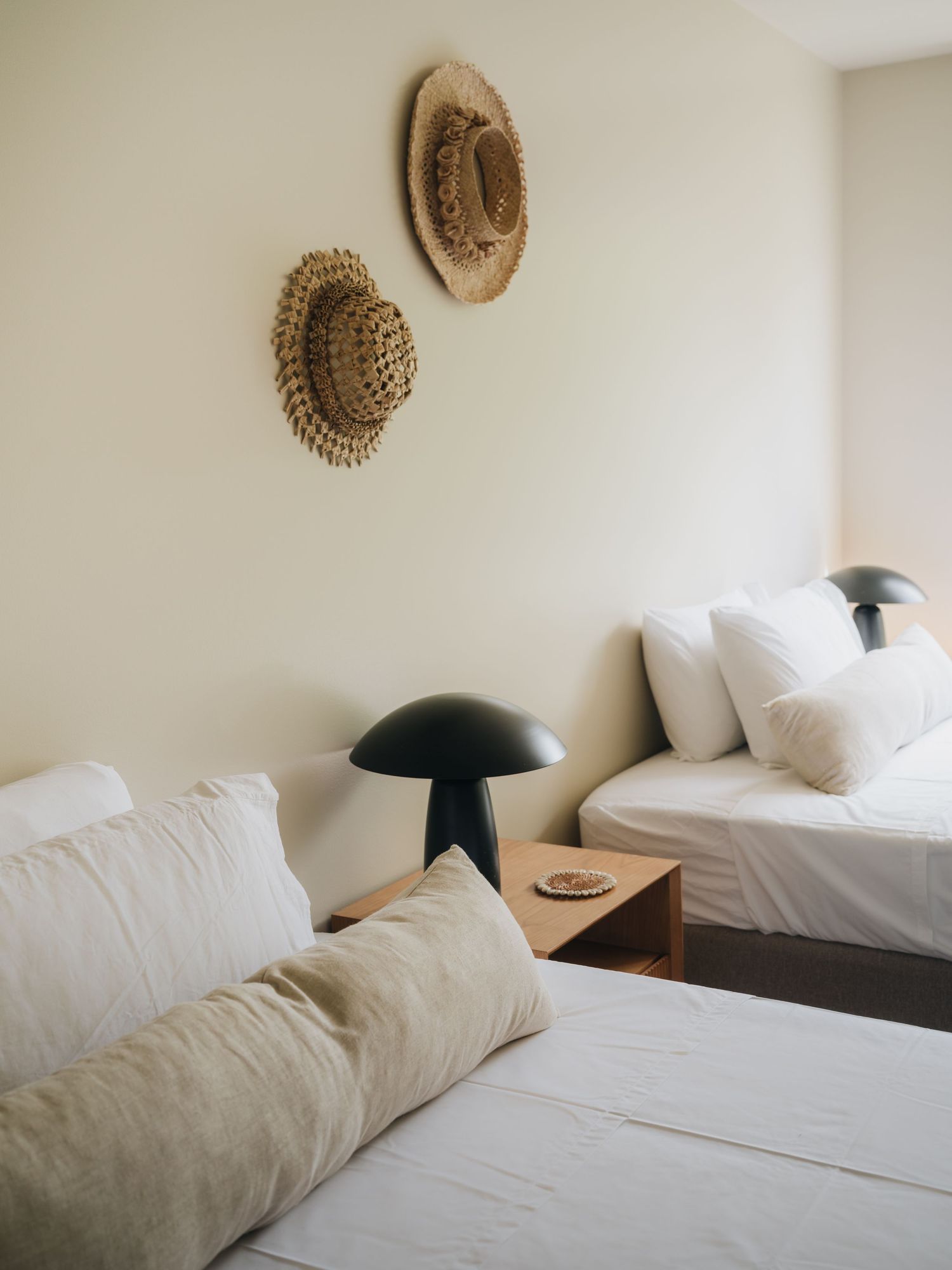Minimalist bedroom with white bedding, mushroom lamp on wood nightstand, and woven straw hats decorating the wall.