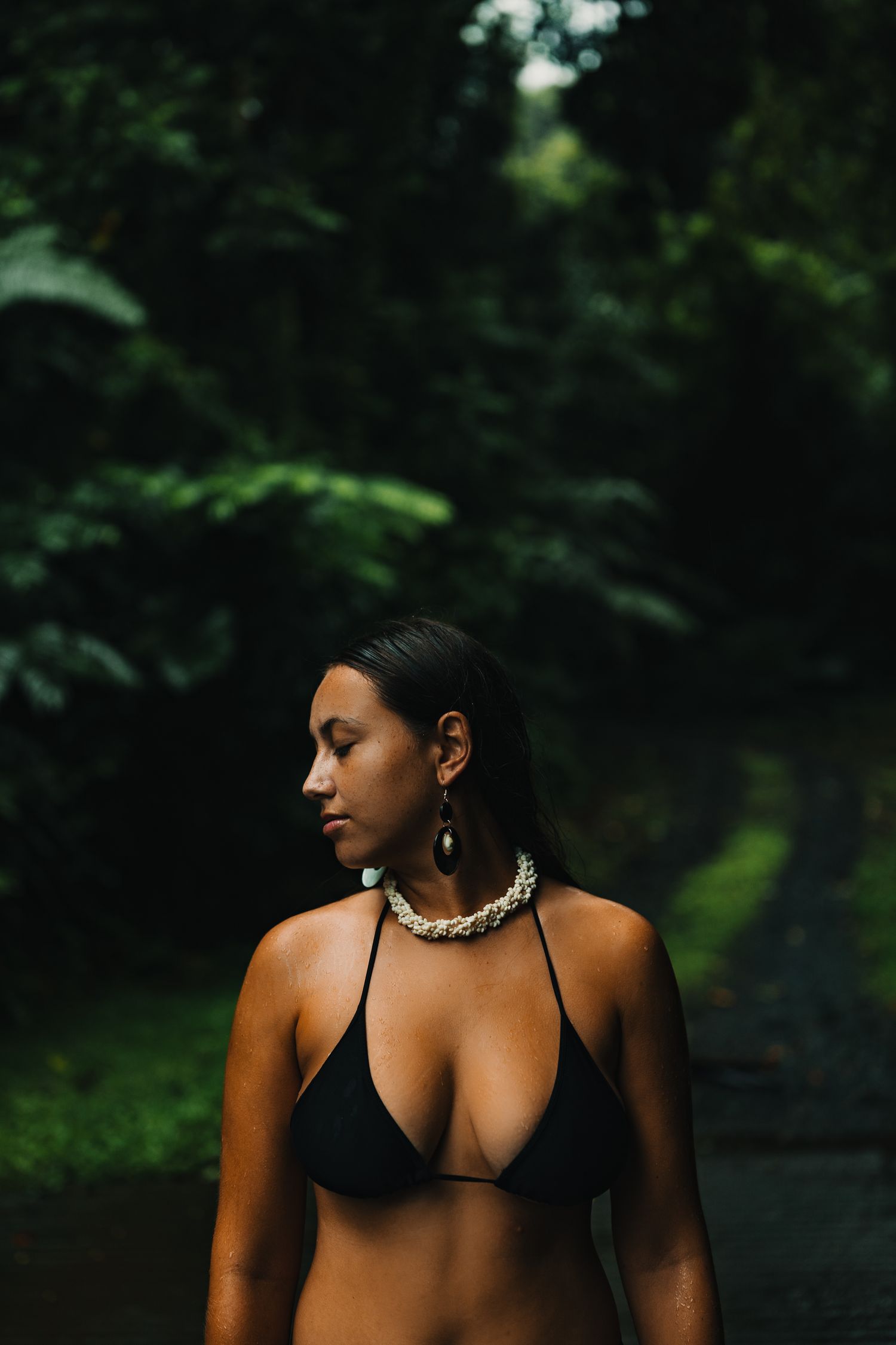 Woman in black bikini and shell jewelry standing in lush tropical forest setting.