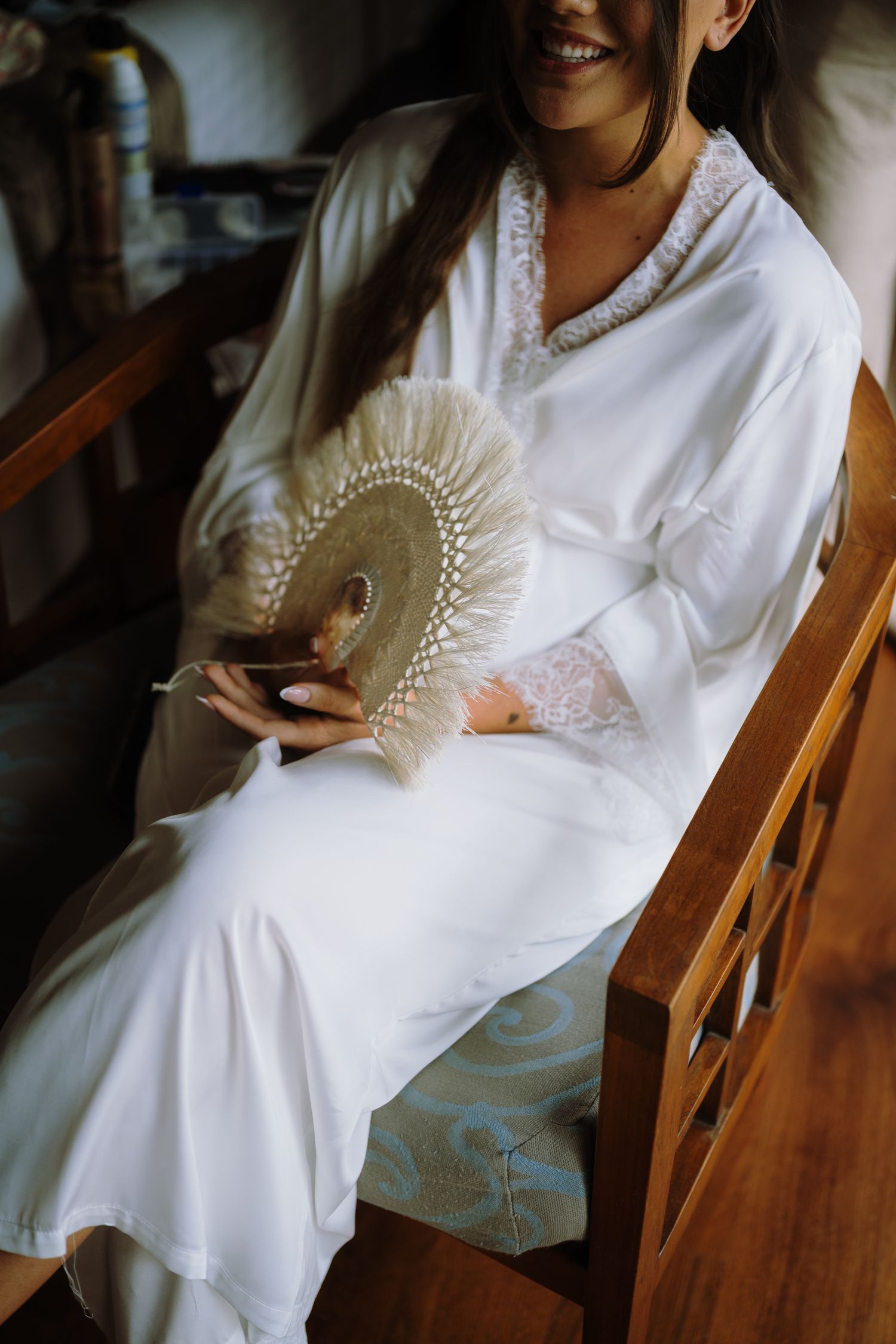 Smiling bride in white robe holding a decorative fan while seated in a wooden chair.