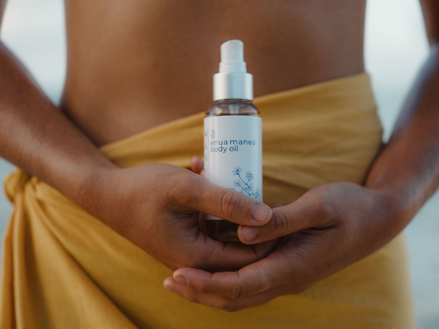 Person holding a spray bottle of mama-mia body oil wrapped in a golden yellow towel outdoors.