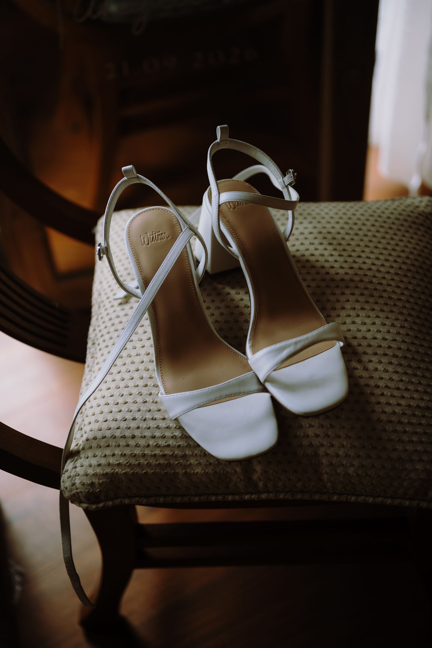 Elegant white strappy high heel sandals resting on a textured upholstered chair, perfect bridal wedding shoes.