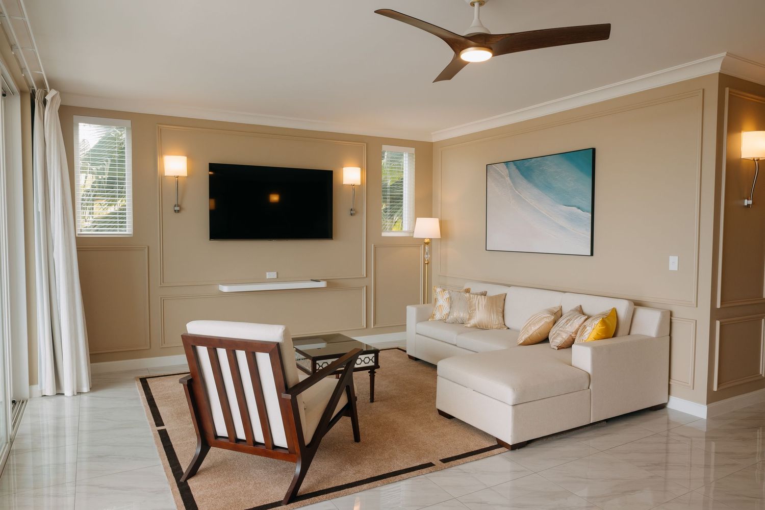 Bright hotel living room with white sectional sofa, ceiling fan, wall-mounted TV, and large windows in a beige interior.