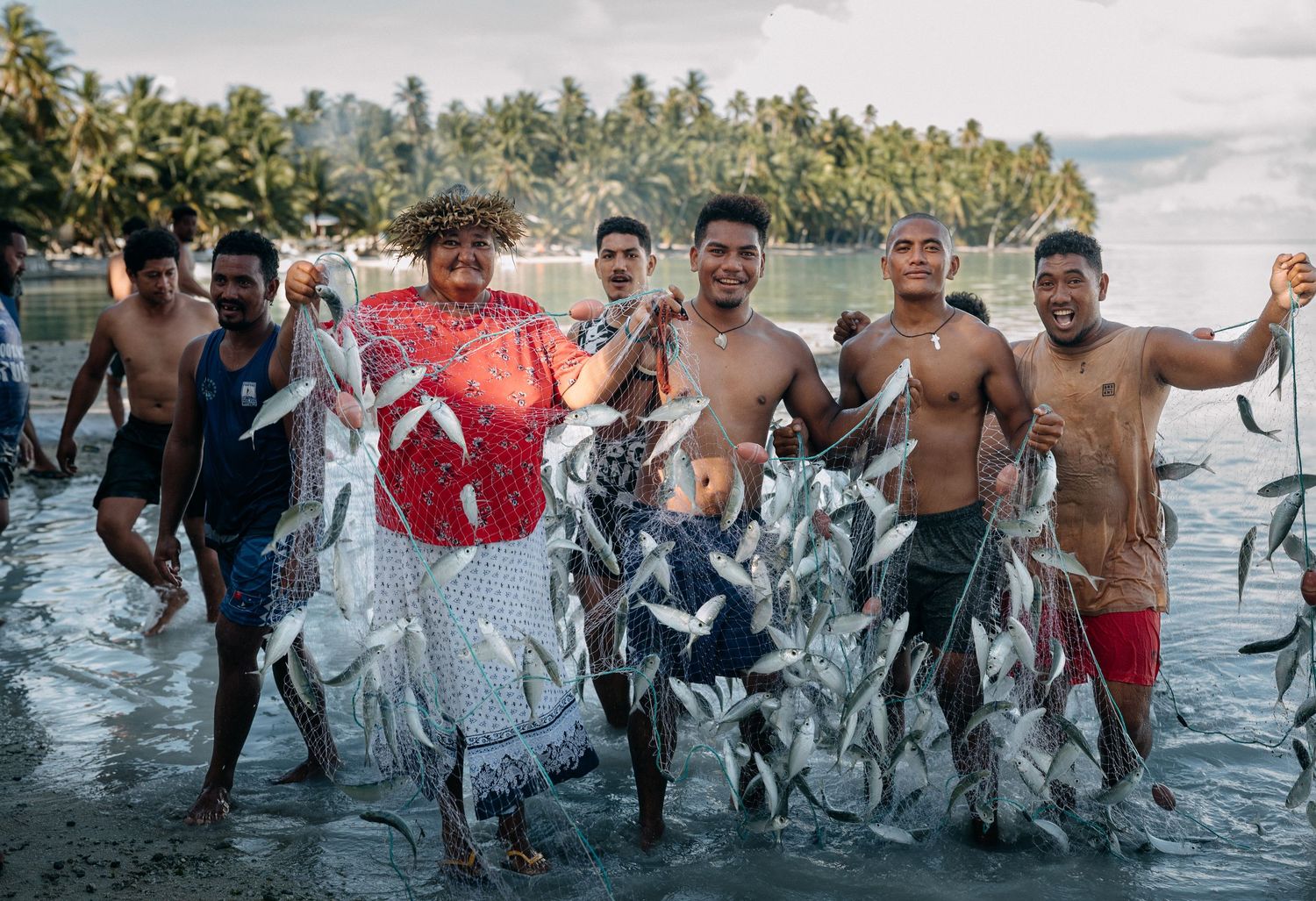 Group of smiling men holding fresh fish catch while standing in shallow coastal waters with palm trees in background.
