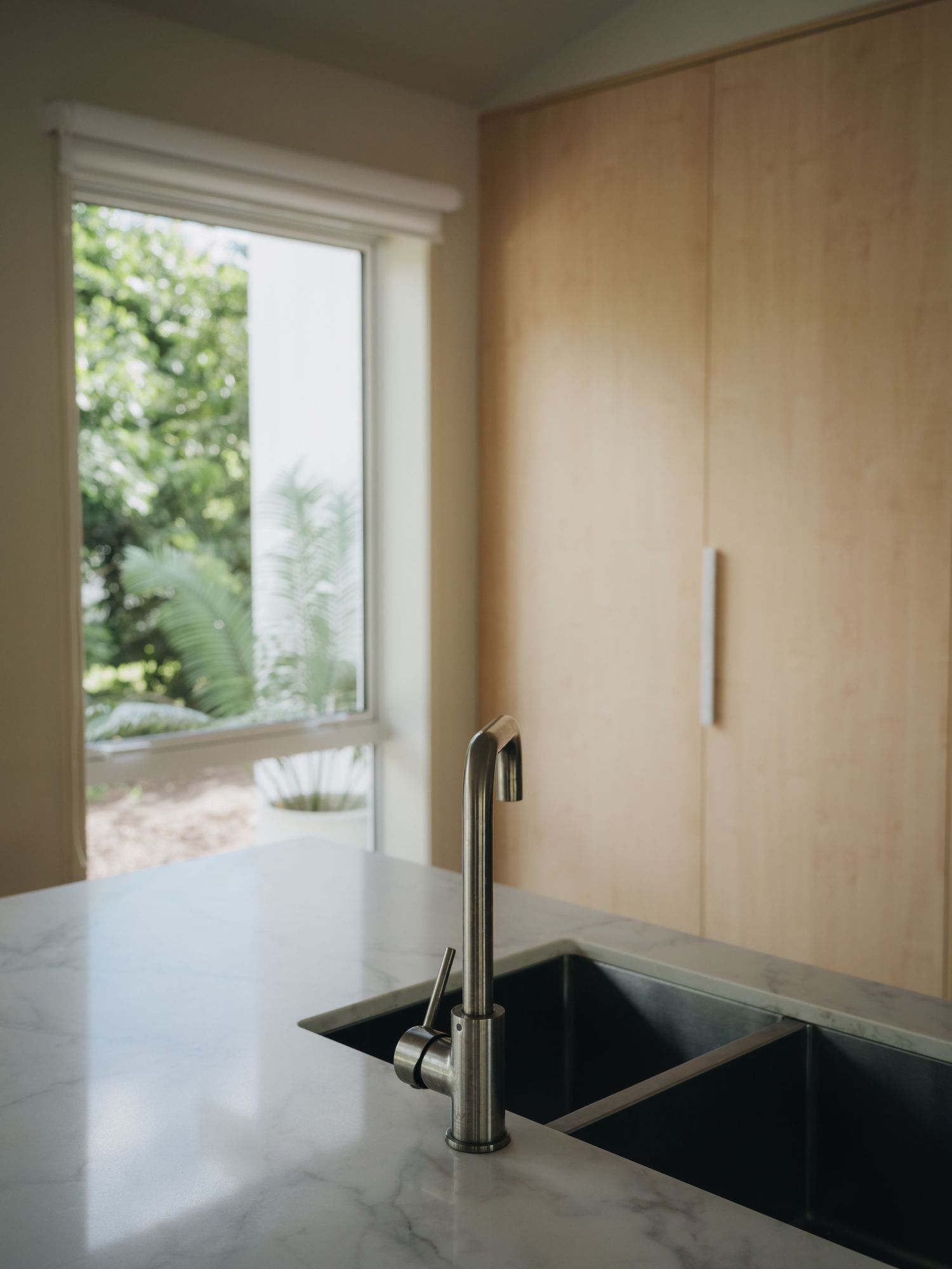 Modern kitchen features marble countertop with stainless steel faucet, undermount sink, and light wood cabinetry near large window.