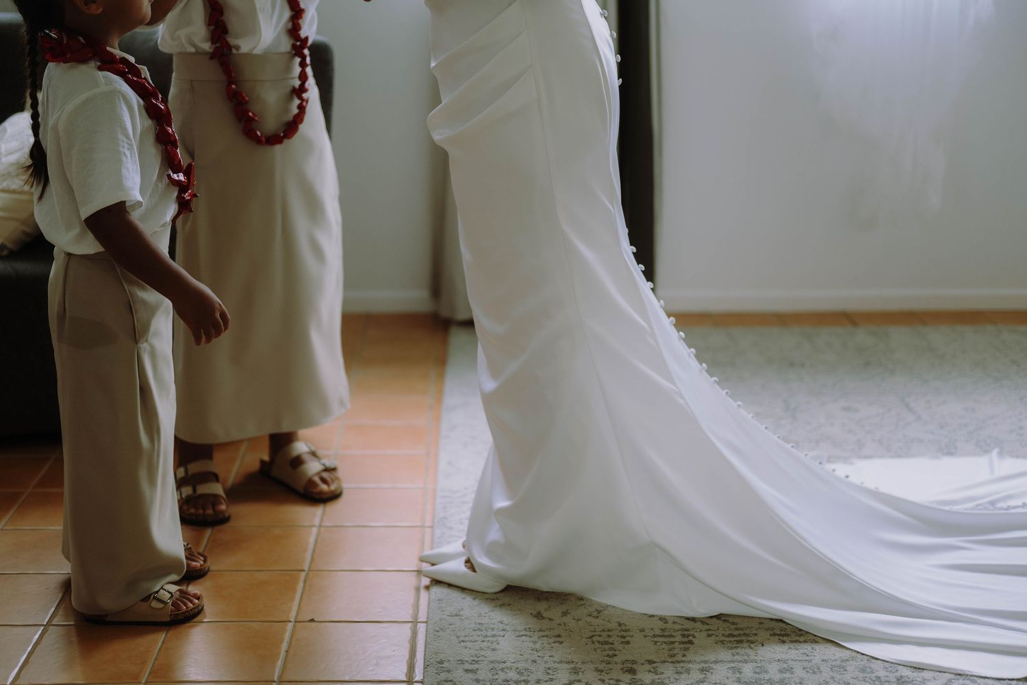 Bride in white wedding gown with train assisted by children in white outfits wearing red leis indoors.