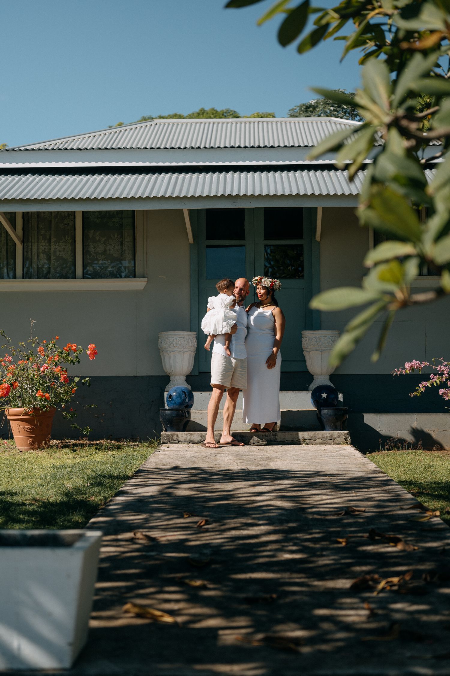 Family in white outfits standing in front of a charming house with lush garden and blue door on a sunny day.