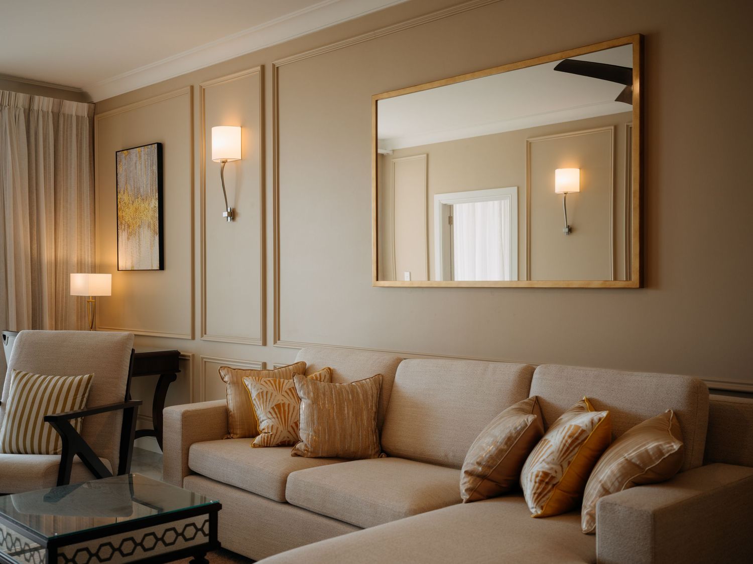 Elegant beige living room with plush sofa, gold-framed mirror, wall sconces, and decorative pillows in warm tones.