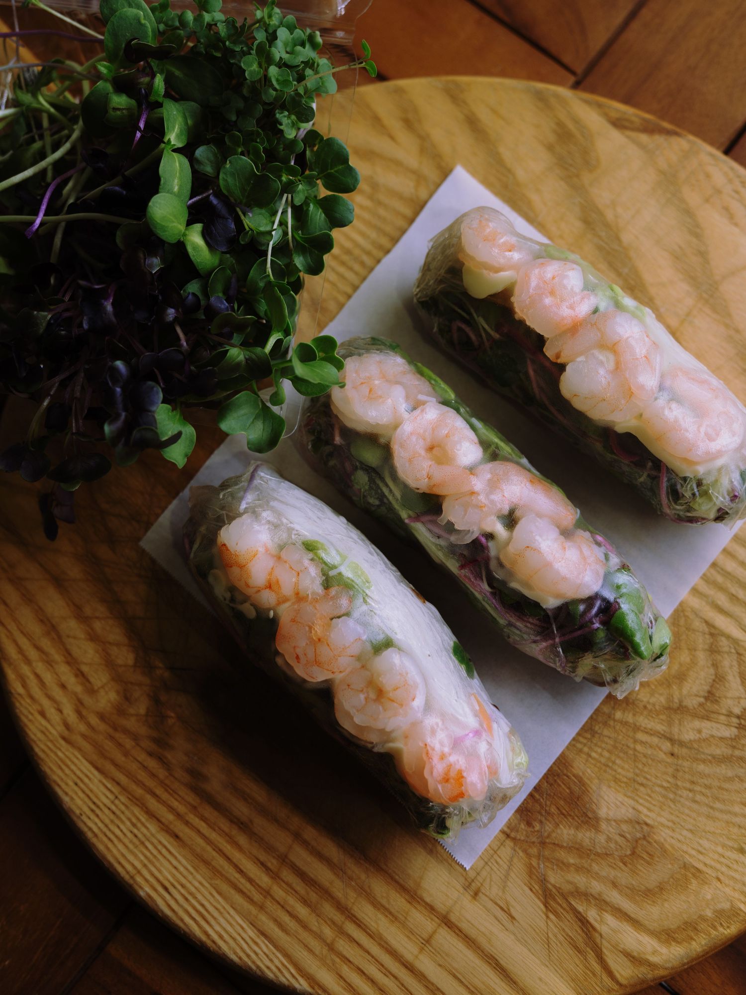 Three fresh shrimp spring rolls with microgreens on a wooden board.