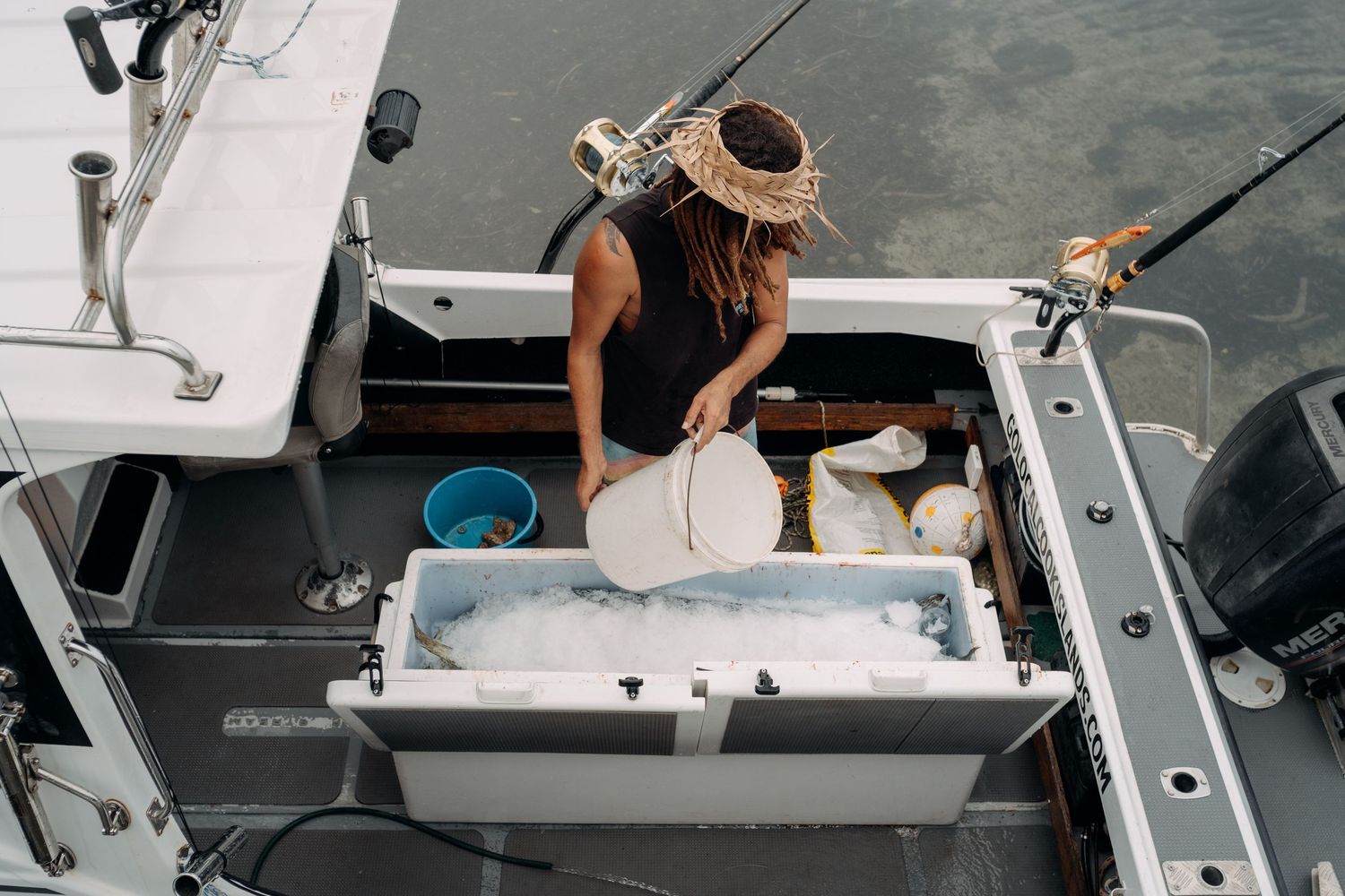 Woman in straw hat sorting fresh catch on ice in a fishing boat cooler, viewed from above on calm water.