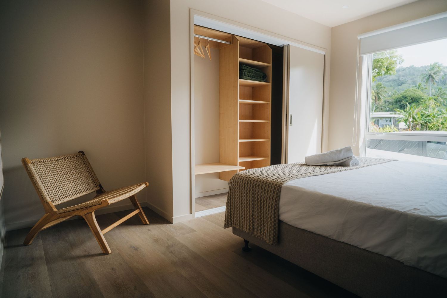 Modern minimalist bedroom with open wardrobe, rattan chair, hardwood floors, and large window overlooking greenery.