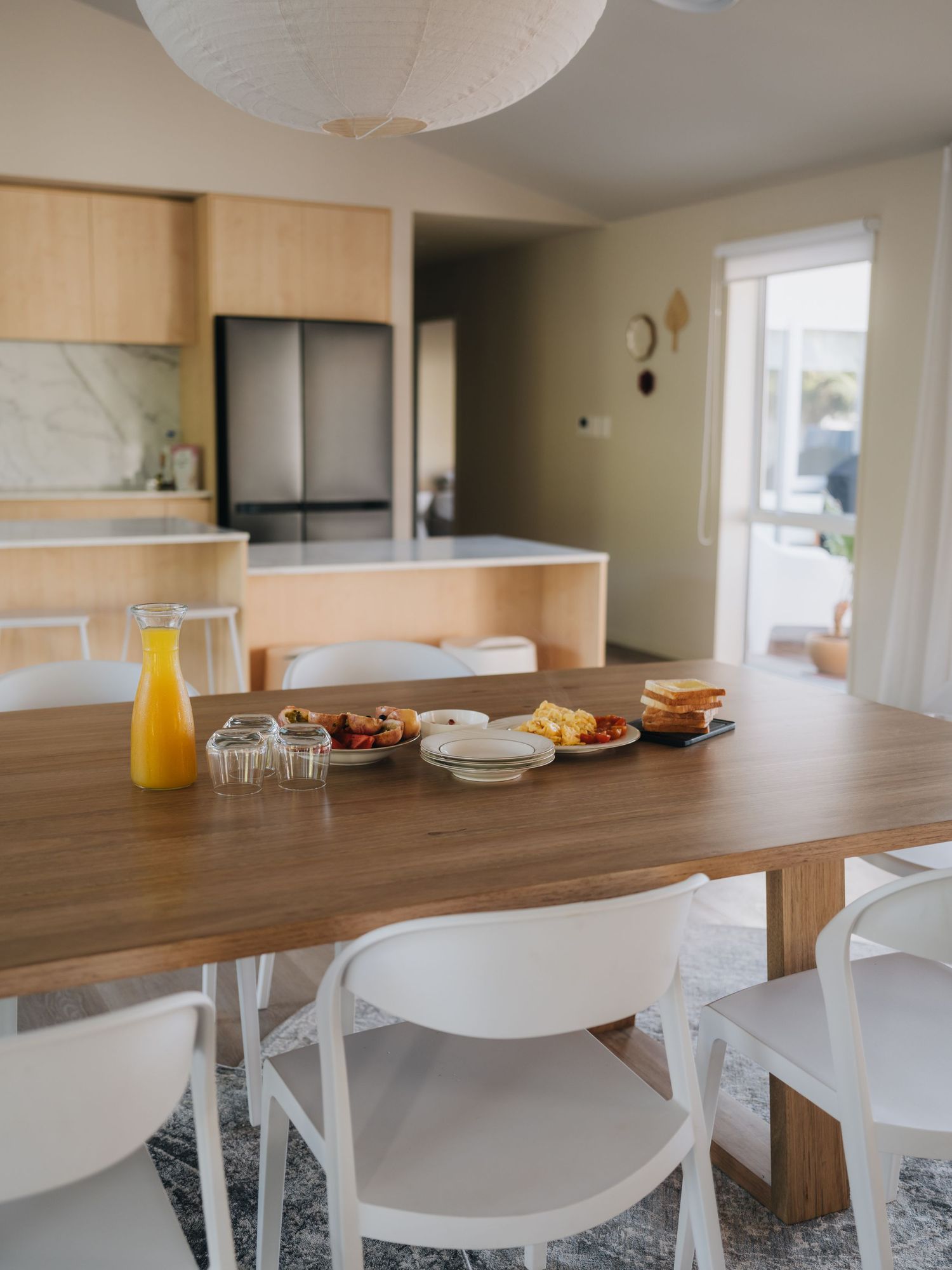 Modern dining area with wooden table, white chairs, orange juice, and open-plan kitchen in background.