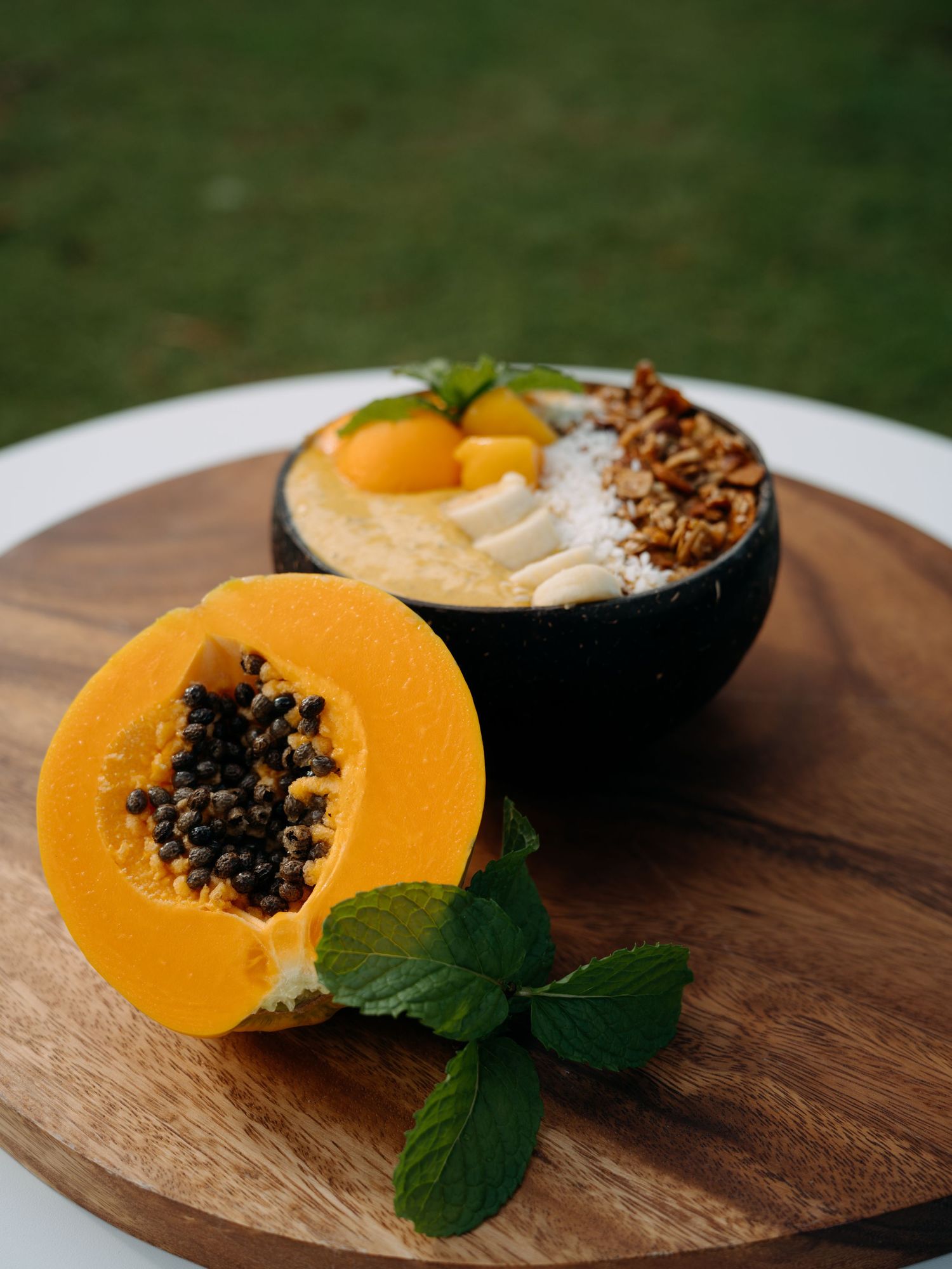 Halved papaya with black seeds on wooden board beside a dark bowl filled with granola, coconut, and tropical fruit toppings.