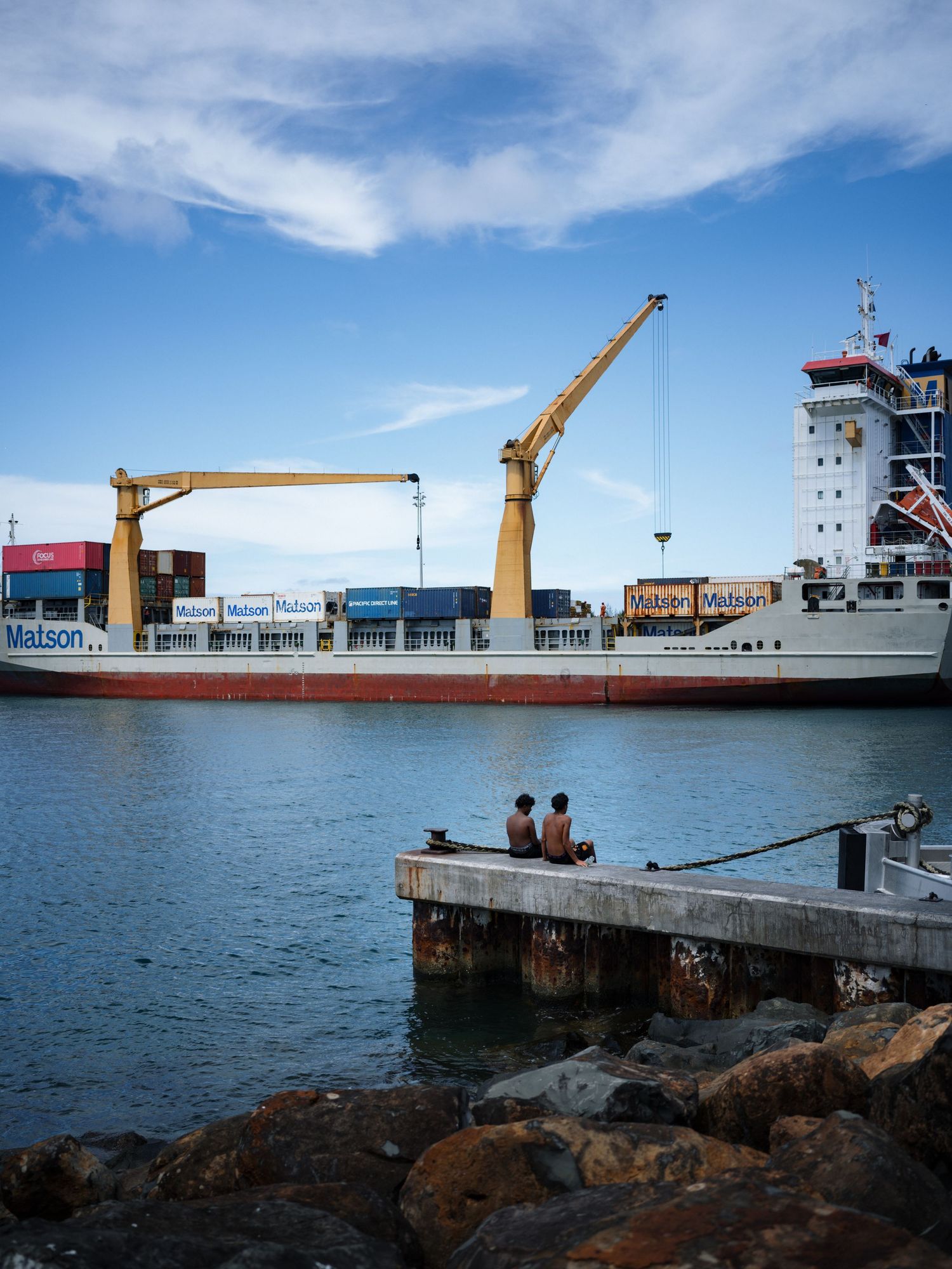 Two people sit on a dock watching a Matson cargo ship with yellow cranes in a busy harbor under blue skies.