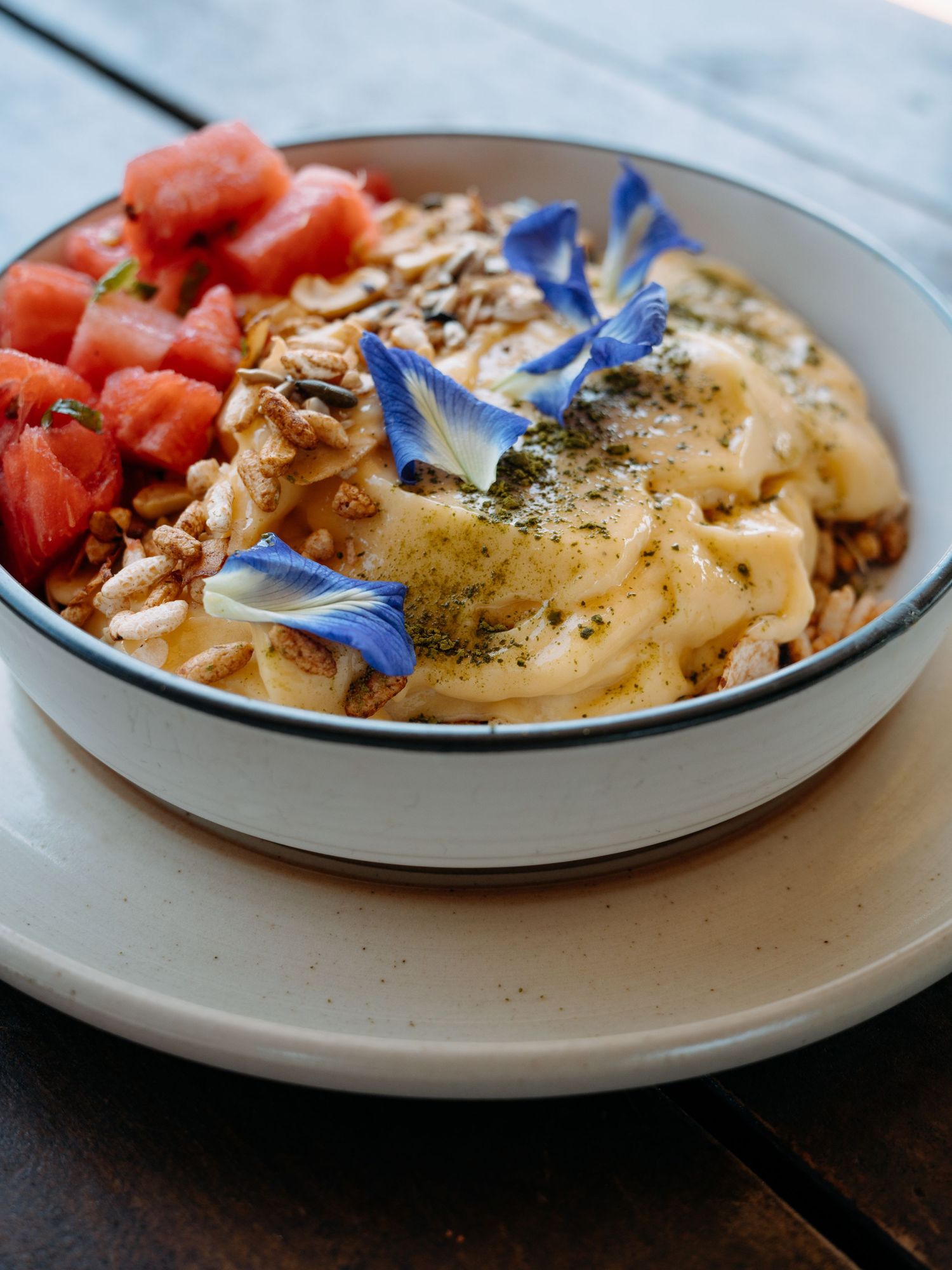 A gourmet bowl with creamy pasta, granola, watermelon chunks, and blue edible flowers on a rustic wooden table.