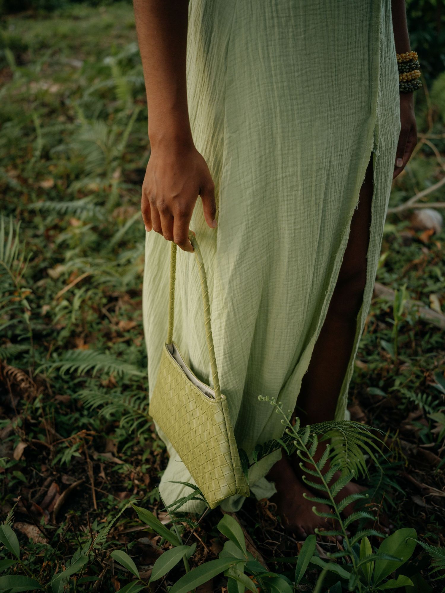 Woman in mint green linen dress holding a woven yellow bag, standing among ferns outdoors.