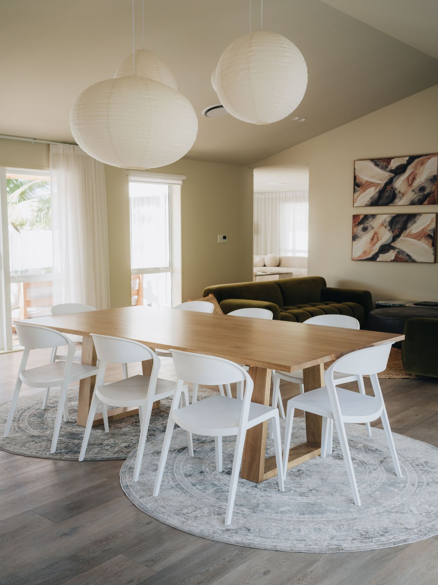 Modern dining room with wooden table, white chairs, paper lantern lights, and a cozy living area in the background.