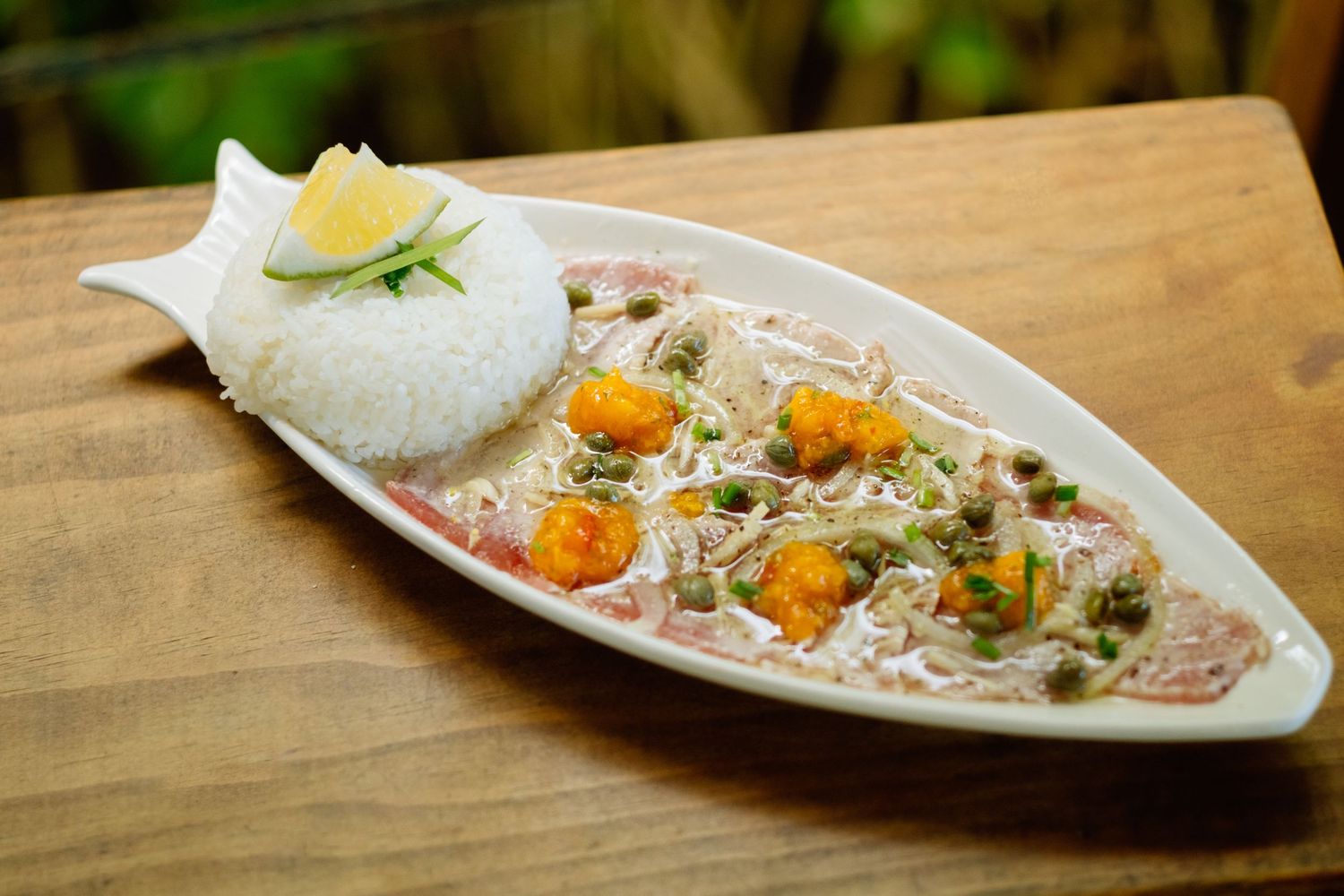 Elegant fish-shaped white plate with creamy seafood dish, rice, capers, shrimp, and lemon garnish on wooden table.