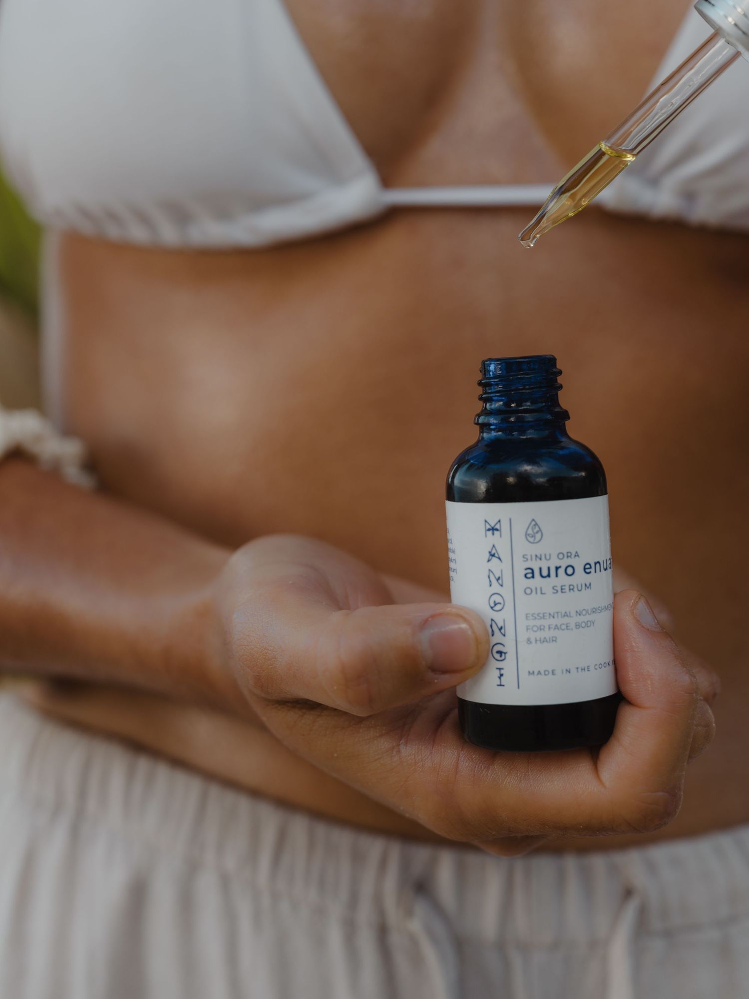 Person holding a dark blue dropper bottle of Auro Enu facial serum, dispensing golden oil drops outdoors.