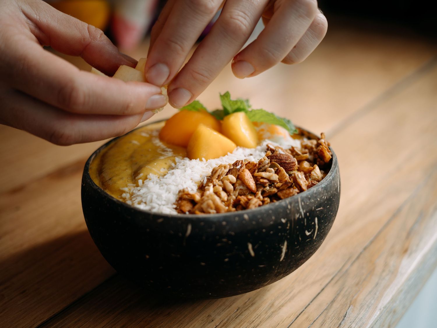 Hands garnishing a smoothie bowl with granola, coconut flakes, and mango chunks in a coconut shell bowl on a wooden table.