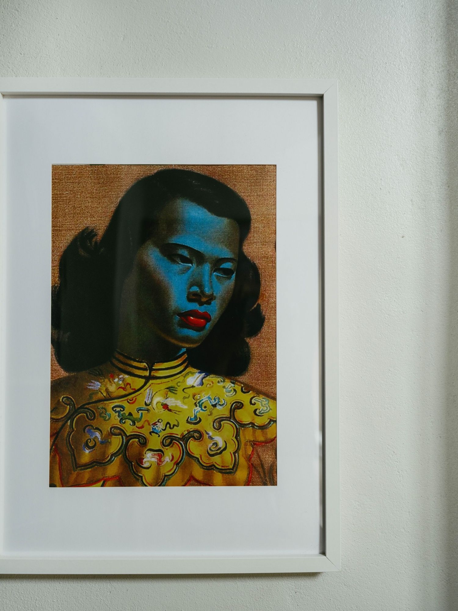 Framed portrait painting of a woman with blue skin wearing a golden embroidered Chinese qipao, displayed on a white wall.