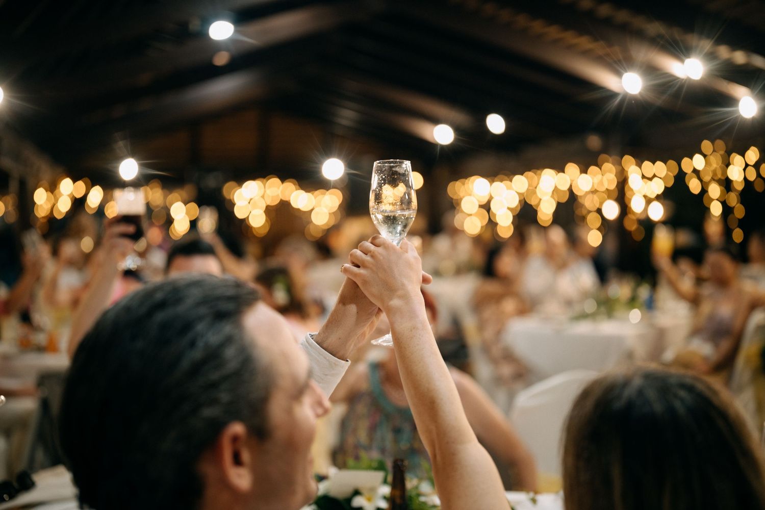 Guests raising champagne glasses in a toast at an elegant evening wedding reception with warm string lights.