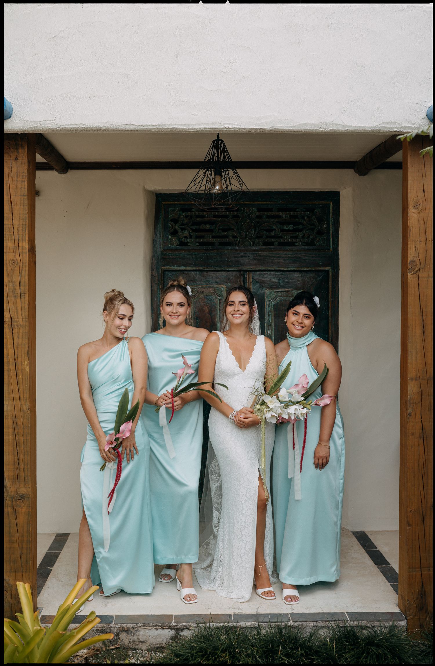 Bride in white lace gown with three bridesmaids in mint green dresses holding tropical bouquets at a wedding venue.