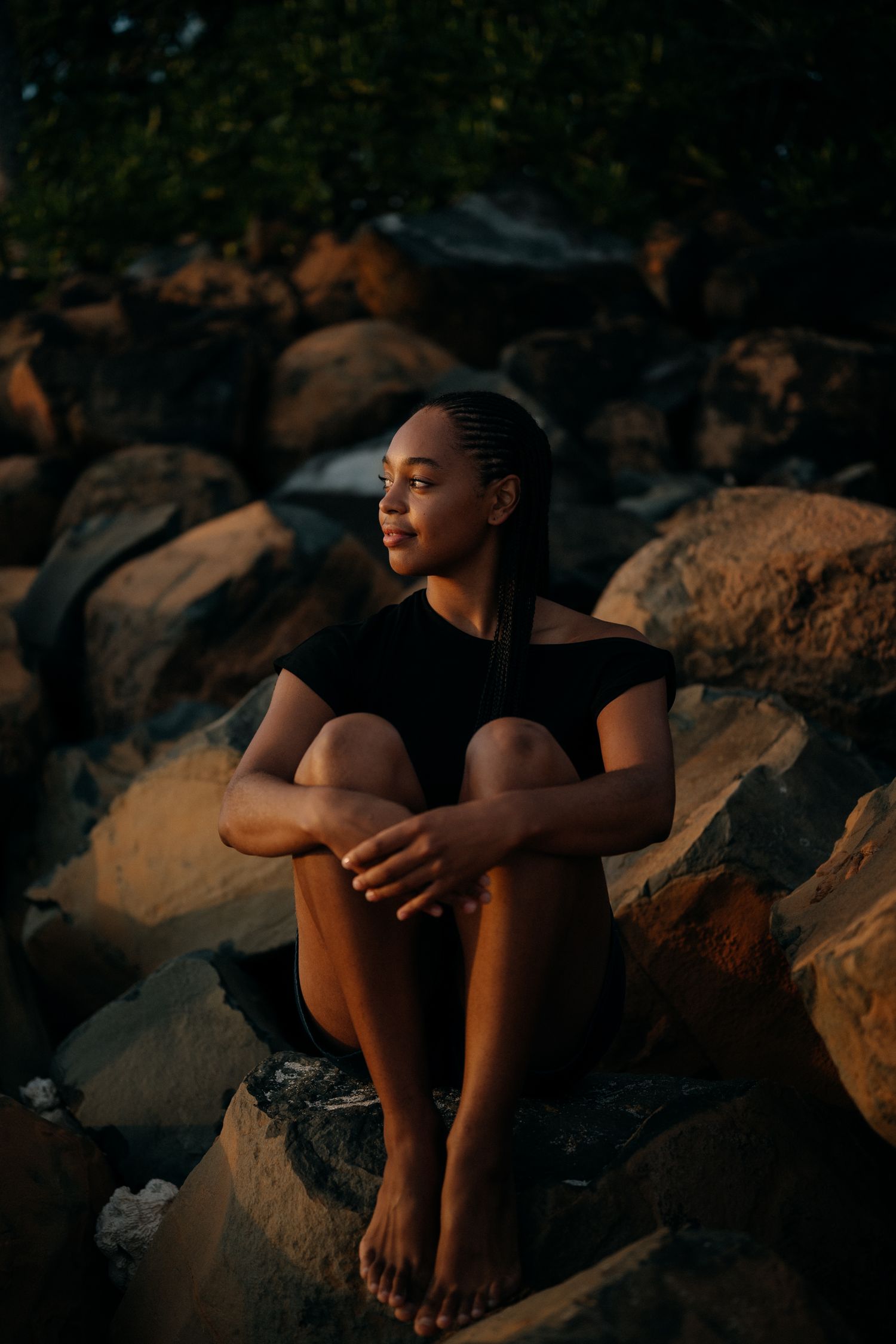 Young Black woman with braids sits barefoot on rocks in golden hour light, wearing a black outfit, gazing peacefully.