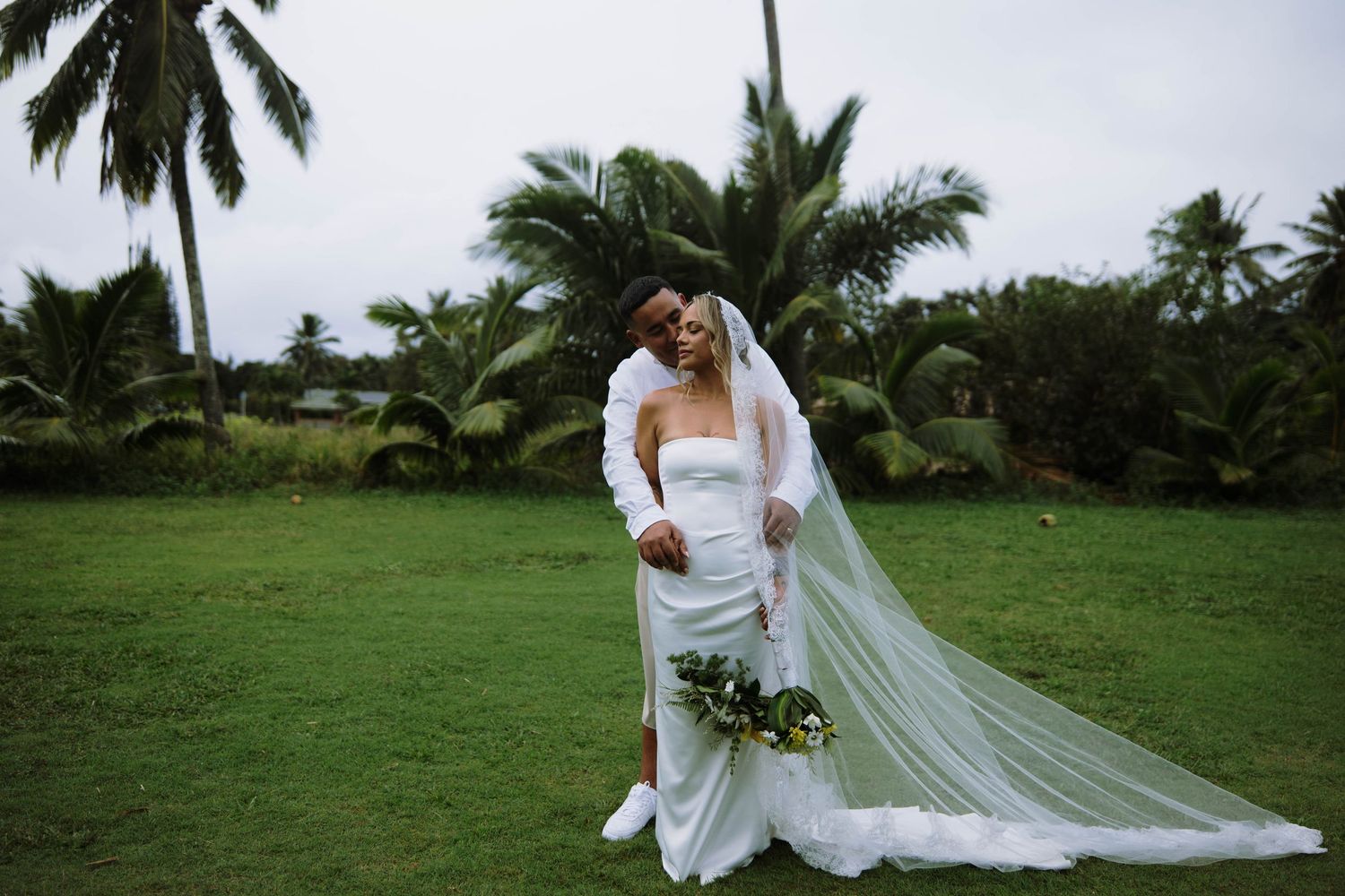 Bride in white strapless gown with flowing veil embraces groom in white suit on lush green lawn with tropical palm trees.