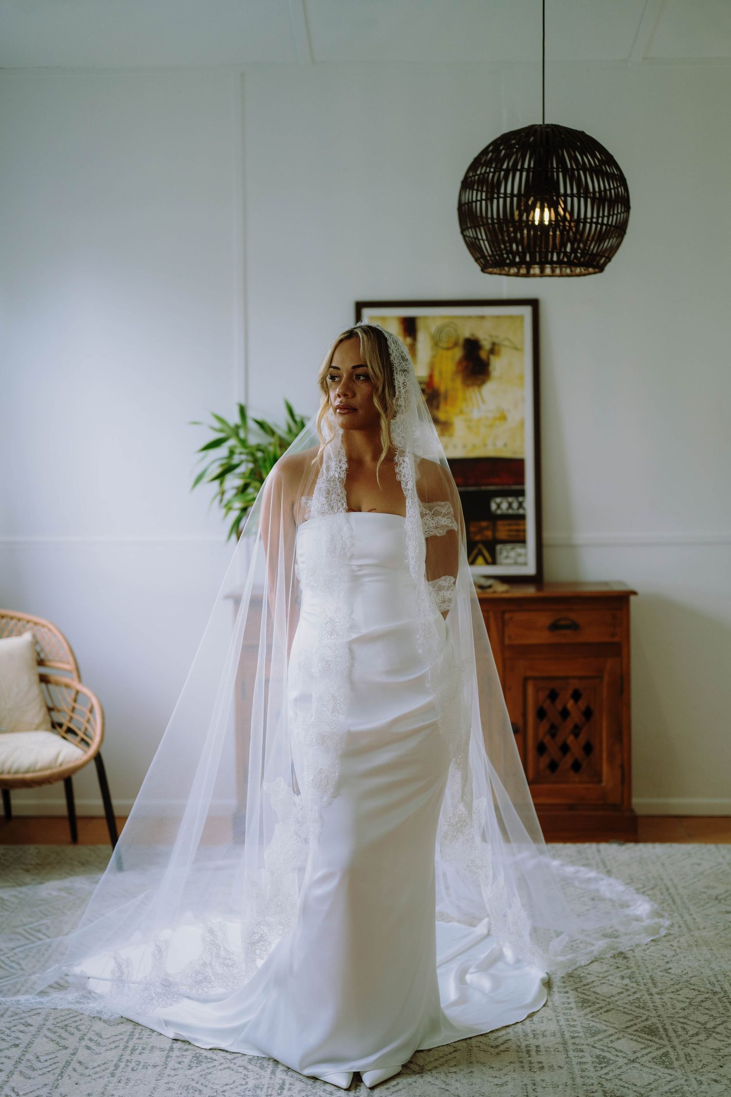 Bride in a white strapless gown with a long lace-trimmed veil stands elegantly in a bright, stylish room.