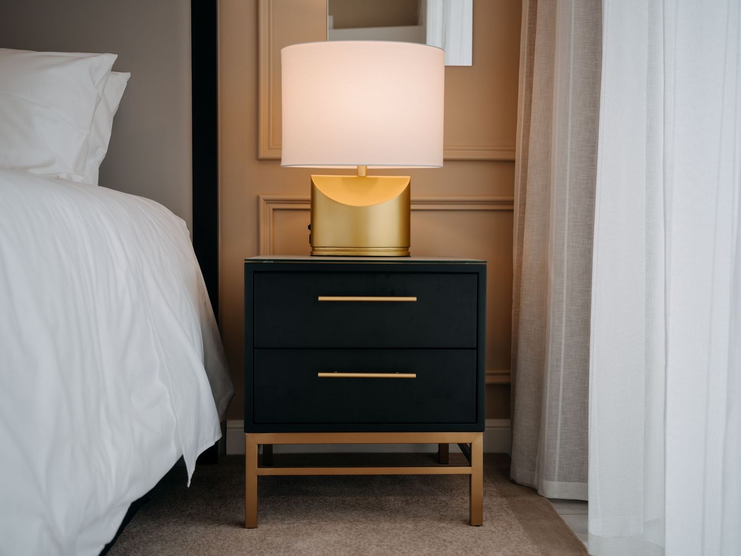 Black and gold nightstand with a gold lamp beside a white bed in an elegant bedroom.
