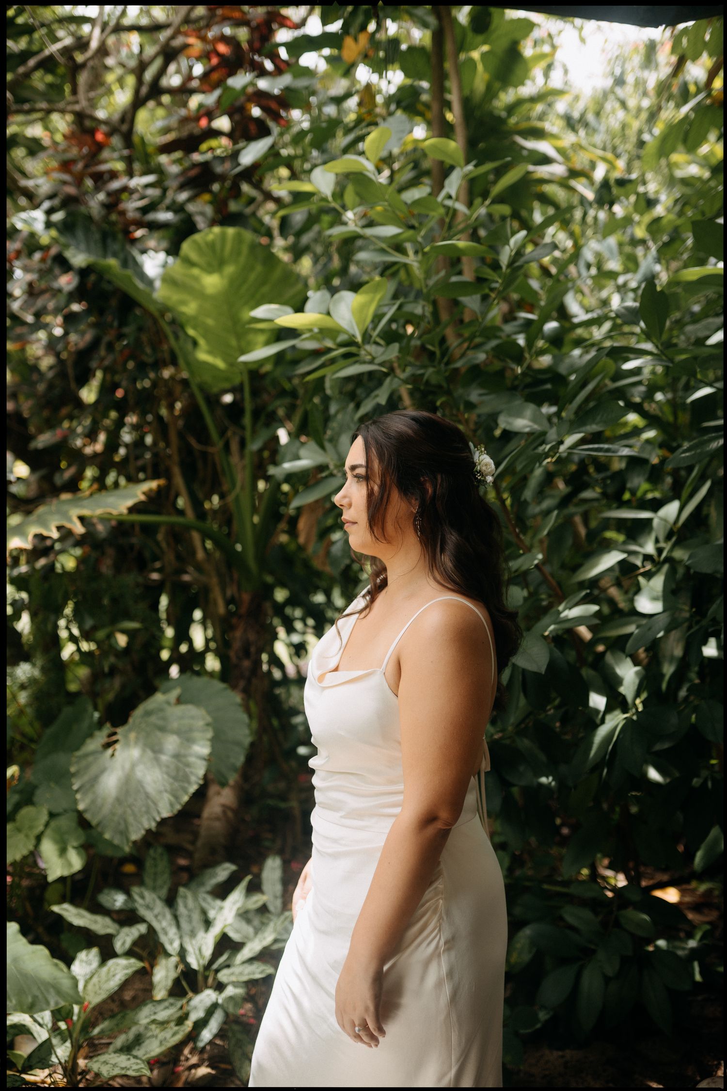Woman in white slip dress standing among lush tropical greenery, looking sideways in a serene outdoor setting.